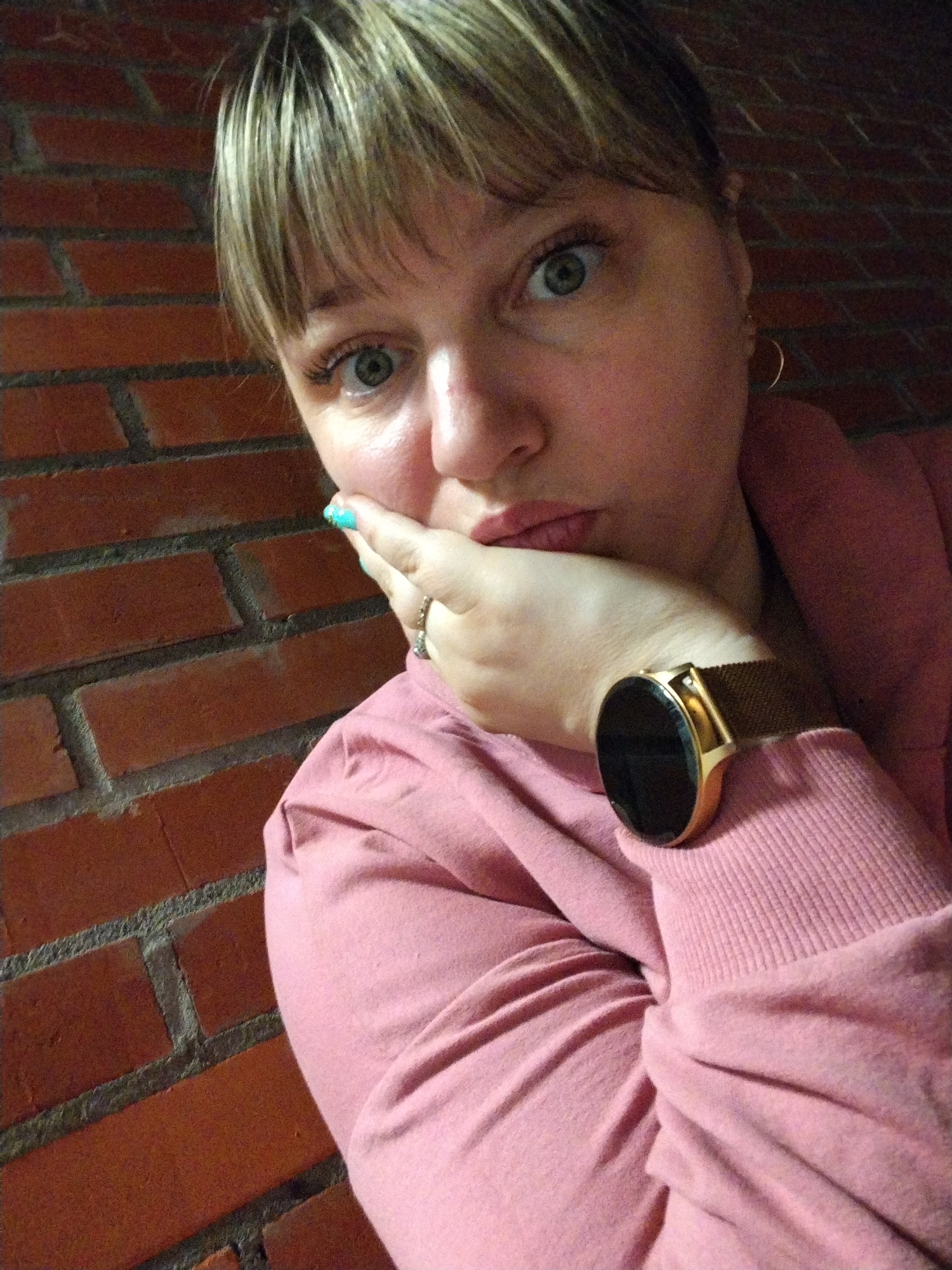An adult woman with light-colored hair, styled with bangs, and striking green eyes, is depicted in a medium close-up, gazing directly at the viewer. She rests her right hand on her chin and lower cheek, revealing light turquoise nail polish on her fingertips and a ring on her ring finger. A gold-toned smartwatch with a dark, round face and a mesh band adorns her left wrist. She is wearing a pink long-sleeved top, possibly a sweatshirt or hoodie, with ribbed cuffs. Small gold hoop earrings are visible in her ears.
The background consists of a reddish-brown brick wall with dark mortar lines, suggesting an indoor or sheltered outdoor setting. The lighting appears somewhat dim, creating soft shadows across the scene. The woman's expression is pensive or contemplative, with a slight pout on her lips. No visible text is present in the image. The location context for this image is Шебунино, Russia.