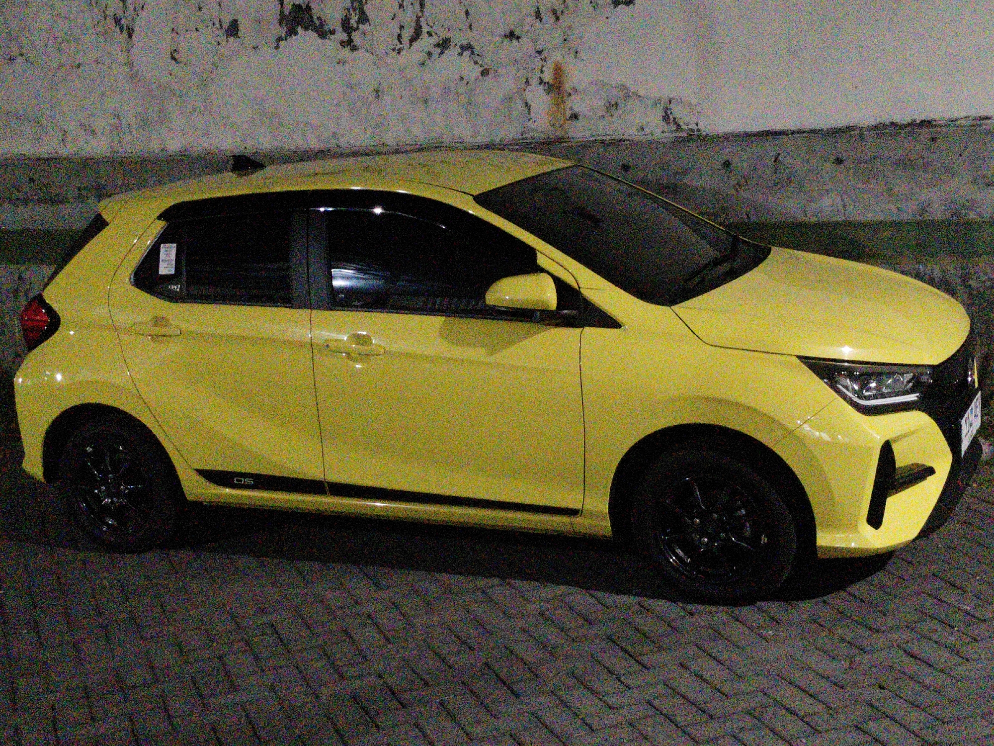 A bright yellow hatchback is parked on a brick-paved surface at night. The car is angled towards the right, with its front end slightly closer to the viewer. It has tinted windows and black wheels. A black stripe runs along the lower side of the car, featuring the text "GS". The background consists of a textured, light-colored wall with dark patches, possibly indicating weathering or damage. The lighting suggests artificial light sources are present, casting shadows on the ground. This scene is located in Semambung, Indonesia.