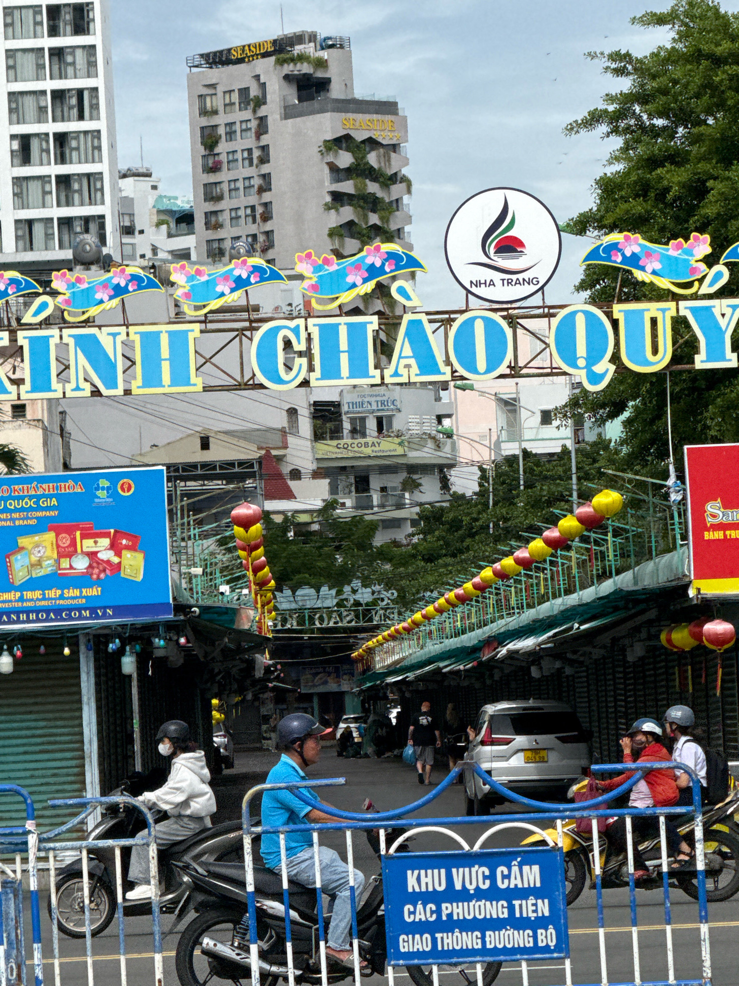 The image captures a bustling street scene in Nha Trang, Vietnam, dominated by a large blue banner with Vietnamese text, likely a welcome or promotional sign. The banner is adorned with decorative blue and pink floral elements. In the background, several buildings of varying heights are visible, including the "SEASIDE" hotel with its distinctive signage and a modern white building with numerous windows. A circular emblem with a stylized logo and "NHA TRANG" text is also prominently displayed on a sign.
The foreground is occupied by a street market with stalls lining the road, indicated by awnings and hanging decorations. A string of red and yellow spherical lanterns stretches across the scene, adding to the festive atmosphere. Several people are present, primarily riding motorcycles. Two individuals are prominently featured on separate motorcycles, both wearing helmets and casual attire. One is on a black motorcycle and the other on a blue motorcycle. Further down the street, other pedestrians and vehicles are visible, suggesting a busy urban environment. A silver SUV is parked in the middle ground. A blue and white striped barrier is in the immediate foreground, with a blue sign displaying Vietnamese text that appears to be a traffic regulation. The overall impression is of a vibrant and active street in a Vietnamese city.