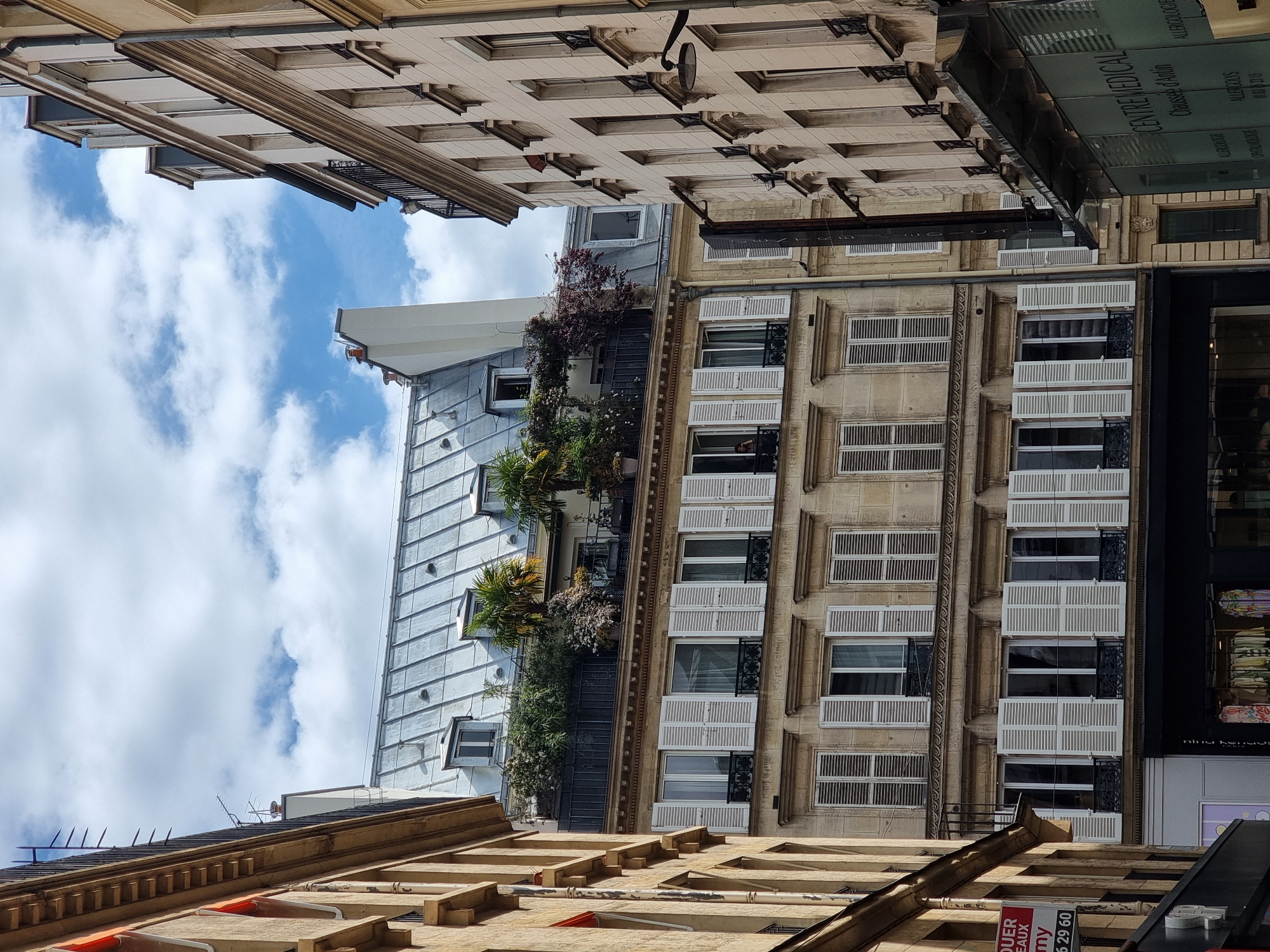The image depicts a street scene in Paris, France, looking upwards at buildings and the sky. The buildings showcase classic Parisian architecture, featuring ornate facades, windows with white shutters, and a metallic roof. Some balconies are adorned with plants. The sky is partly cloudy. The photograph is taken from a low angle, possibly street level, looking up towards the buildings and the sky above.
