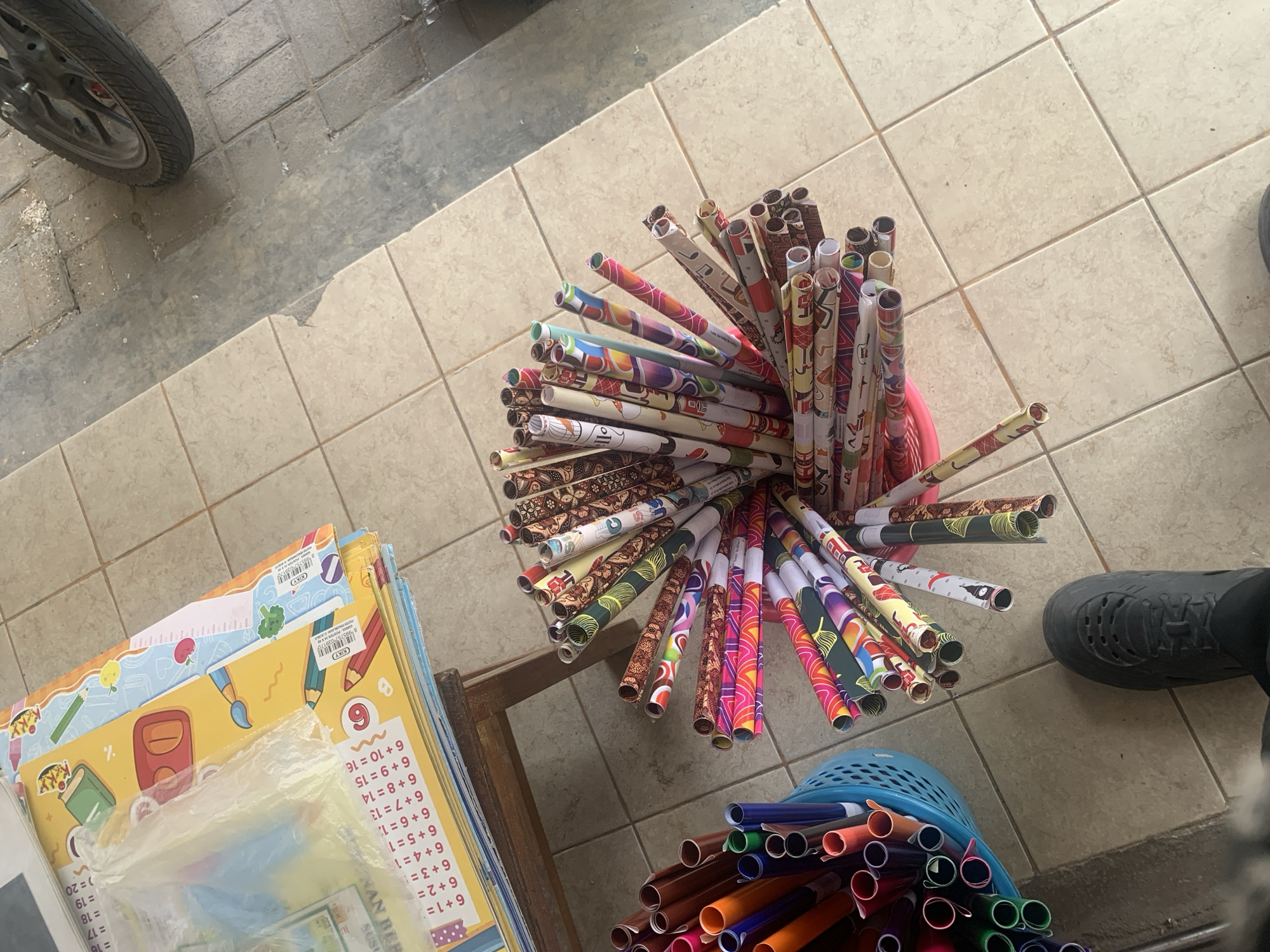 In Palembang, Indonesia, a street-level view captures a display of rolled papers, likely for sale. A collection of colorful, patterned paper rolls is arranged in a pink container, with some scattered around it. Nearby, a blue plastic basket overflows with individual paper tubes of various colors, including orange, purple, blue, and green. In the background, stacks of educational posters, featuring numbers and illustrations of school supplies like pencils and paintbrushes, are piled on a wooden surface. The tiled floor suggests an outdoor or semi-outdoor market setting. A portion of a motorcycle wheel is visible in the upper right corner. A person's foot, clad in a black sneaker, is positioned in the lower center of the frame, indicating a potential shopper. The overall scene depicts a vibrant, commercial environment focused on art and educational supplies.