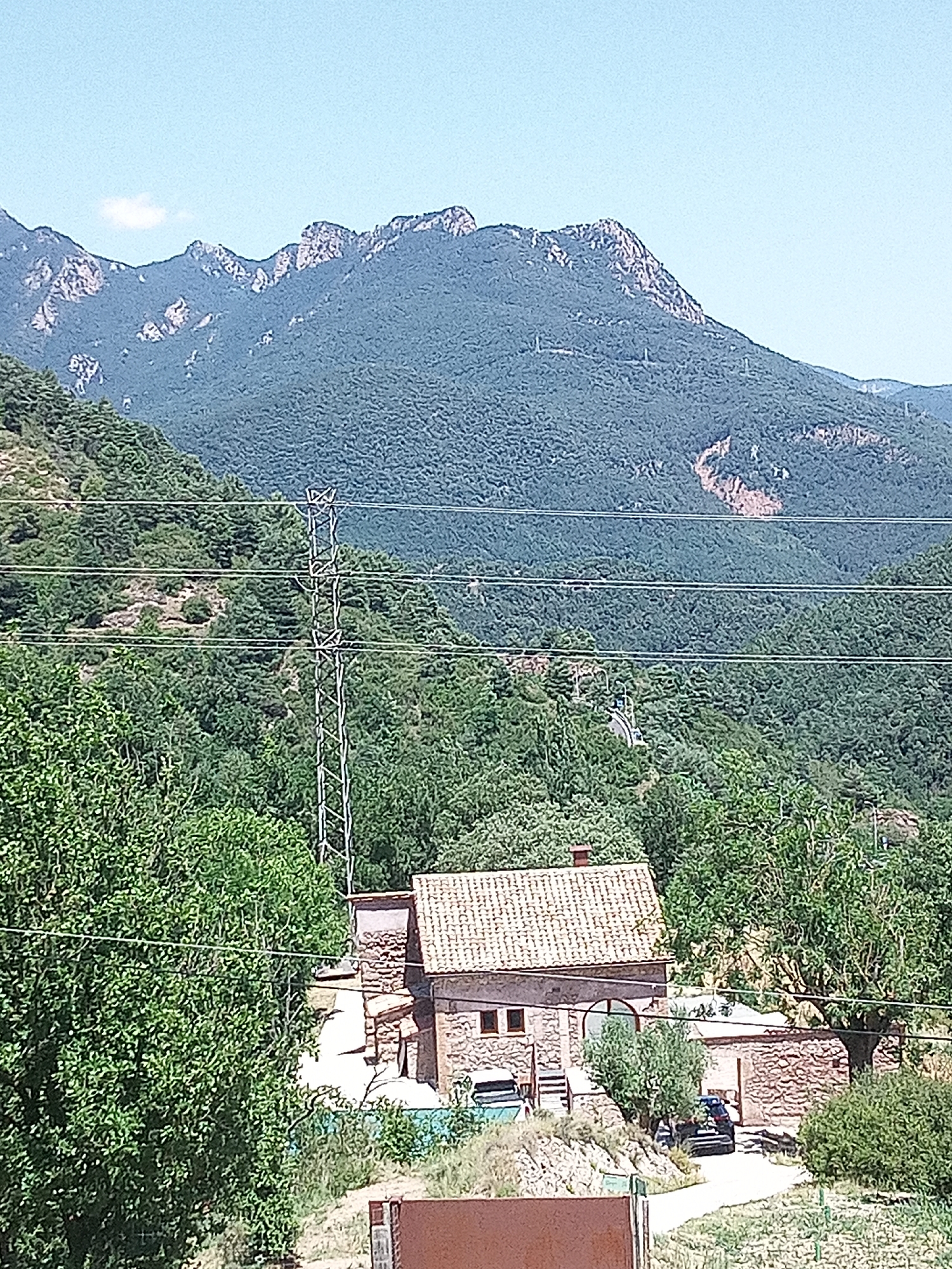 The image depicts a rural scene with mountains in the background and a house in the foreground. The mountains are covered in dense green vegetation and have rocky peaks. A few power lines are strung across the landscape, supported by tall metal towers. In the middle ground, a stone house with a tiled roof is visible, surrounded by trees. A dark-colored car is parked on a dirt road in front of the house. The sky is clear and blue, with a single white cloud. The overall impression is one of a serene, natural setting. Given the context of Berga, Spain, this scene likely represents a mountainous area in or around the city, possibly a rural property or a hiking area.