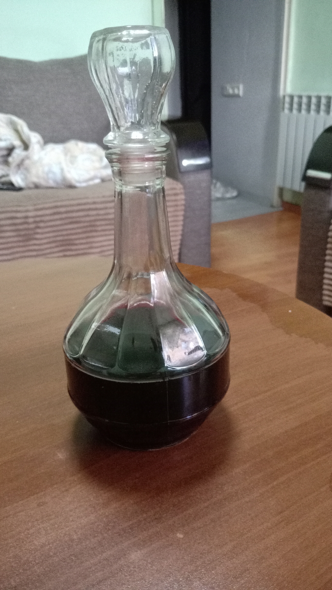 A glass decanter containing a dark liquid sits on a wooden surface. The decanter has a rounded bottom with a black base, a clear glass body with visible liquid fill, and a clear glass stopper. In the background, there is a couch partially covered with a light-colored blanket, along with a radiator and a section of a wall. The location appears to be indoors, possibly a residential setting. The city and specific location are unknown.