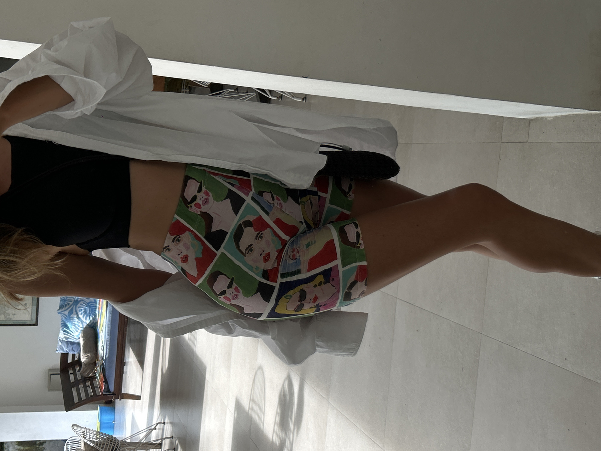 A woman is standing indoors in Pererenan, Indonesia, wearing a black top, patterned shorts featuring faces, and a white button-down shirt. She is positioned on a light-colored tiled floor. In the background, there is a daybed with blue and patterned pillows. Sunlight is streaming into the room. A white chair is visible in the distance.