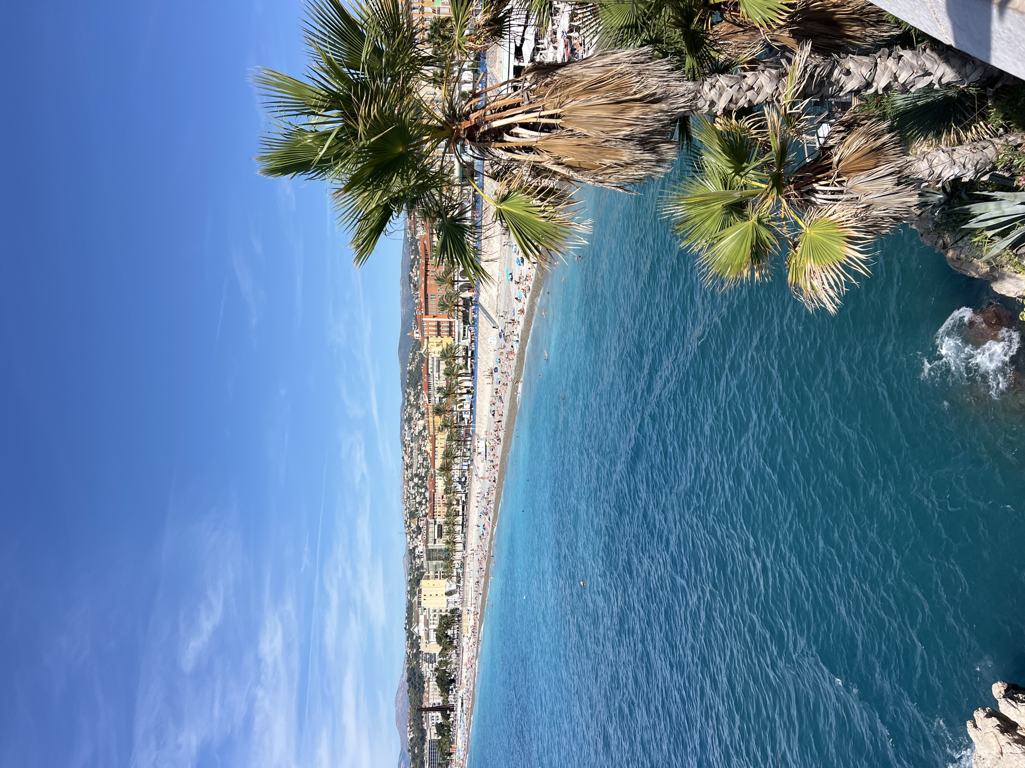 This is a high-angle, eye-level, outdoor, daytime, full shot of the Promenade des Anglais in Nice, France, on a sunny day. The image captures the vibrant blue waters of the Mediterranean Sea in the foreground, with gentle waves lapping against the rocky coastline on the left. A crowded beach lines the shore, with numerous people sunbathing and swimming. Behind the beach, a bustling promenade is visible, lined with palm trees and buildings in various shades of yellow, orange, and cream. The sky is a clear, bright blue with a few wispy white clouds. In the foreground, two palm trees frame the scene, their fronds casting shadows on the water.