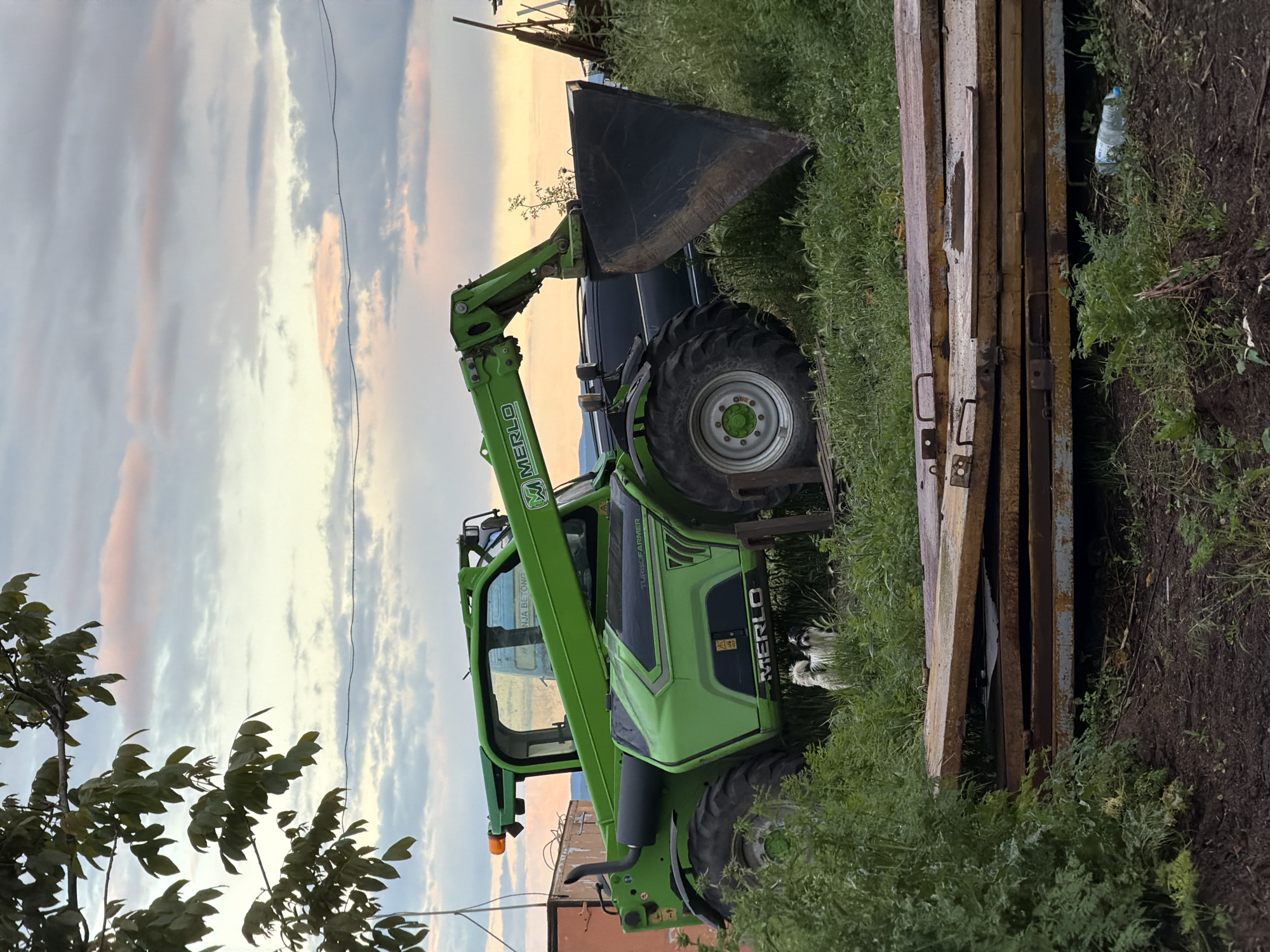 The image depicts a Merlo Turbofarmer telescopic handler parked outdoors near Cherni Vrah, Bulgaria. The machine is green and black. It has a large front bucket. The handler is parked on a grassy area beside metal gates. The sky is cloudy, with hints of sunset. A utility wire is visible above. Foliage is present in the foreground and background.