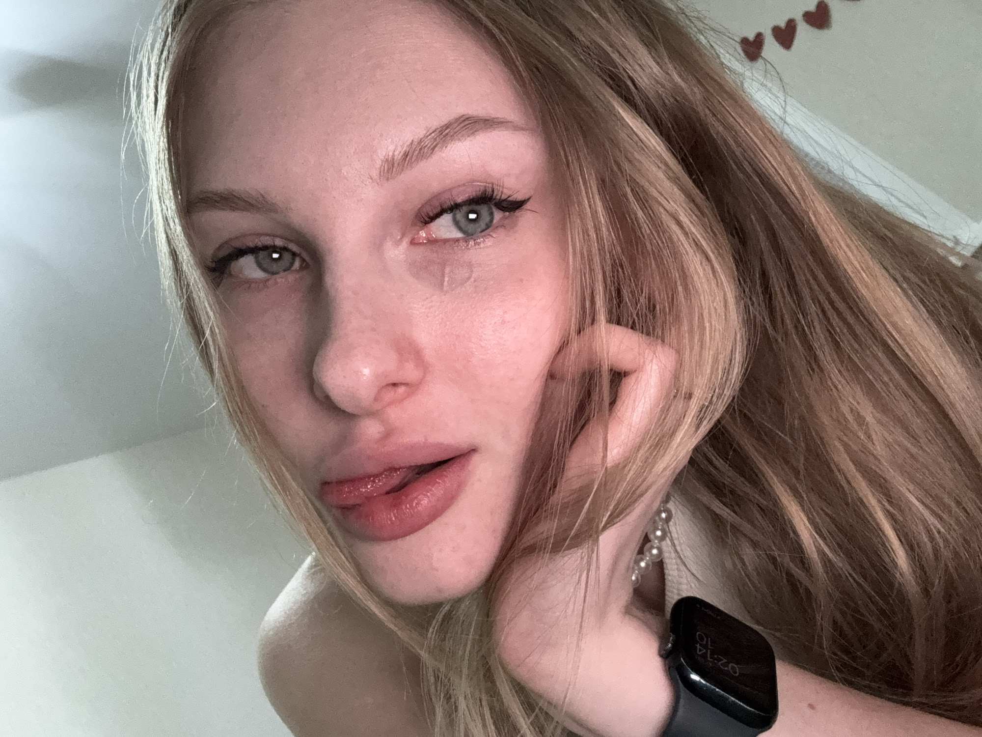 A close-up, overhead shot features a young Caucasian woman with blonde hair and blue eyes. Her head is tilted to the right, with her right cheek resting on her hand, which is adorned with a smartwatch. Her mouth is slightly open, with her tongue visible. She is wearing a pearl necklace. The background appears to be a plain wall, with a decorative element of small red hearts visible on the right side. There is no indication of the city of Perm, Russia, or any other specific location.