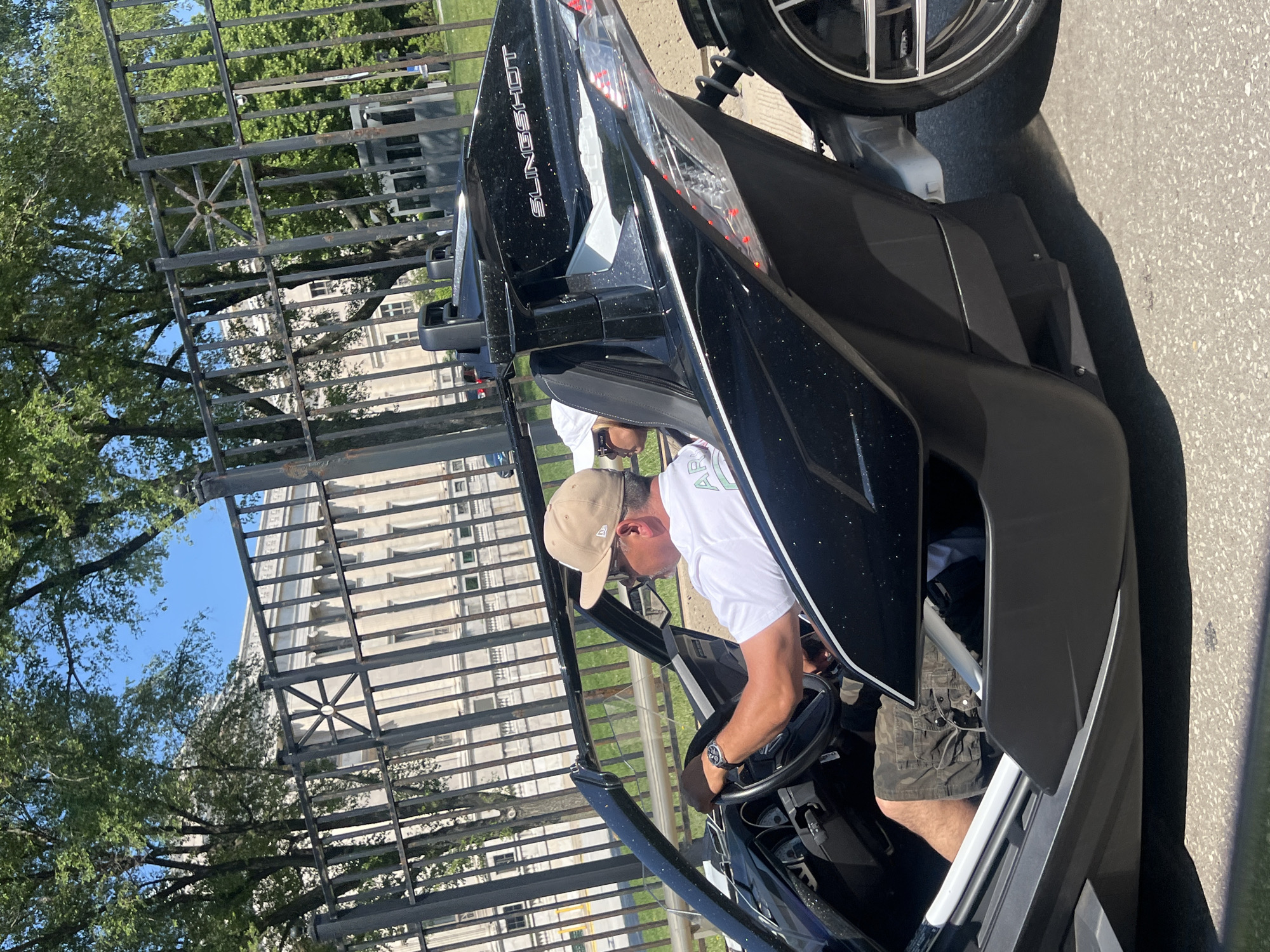 A man is sitting in the driver's seat of a black Polaris Slingshot parked on a paved surface. He is wearing a white t-shirt, tan hat, and camouflage shorts, and he is looking at the steering wheel. The car is parked next to a black metal fence, and behind the fence, there are trees and a tall building. The image appears to be taken outdoors on a sunny day in a city.