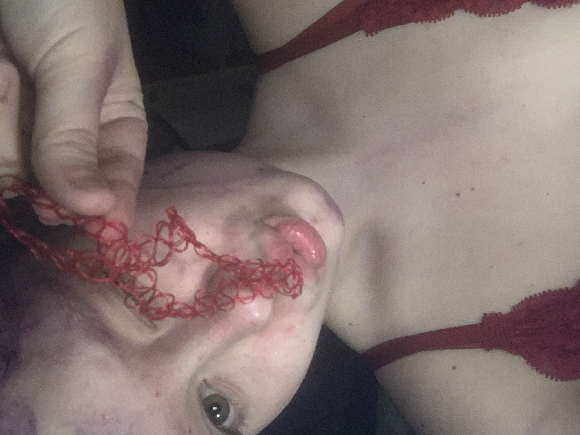 The image captures a close-up, rotated view of a person, likely taken indoors in Vsevolozhsk, Russia, under dim lighting conditions. The subject is light-skinned, with their head and upper torso visible. They are wearing a red lace bra, the strap and upper trim of which are discernible.
A hand, appearing to belong to the subject, holds a red, intricate string-like object, possibly a piece of lace or a decorative string, close to the person's face. The person's mouth is slightly open, revealing their tongue, while their left eye is open and looking directly at the viewer.
Notable details include several purplish marks on the person's neck and extending onto their face, which could indicate bruising or be part of a deliberate aesthetic like makeup. Additionally, small reddish spots or moles are scattered across their skin. No text is visible in the media file. The overall impression is intimate and somewhat enigmatic, presented from an unconventional angle.