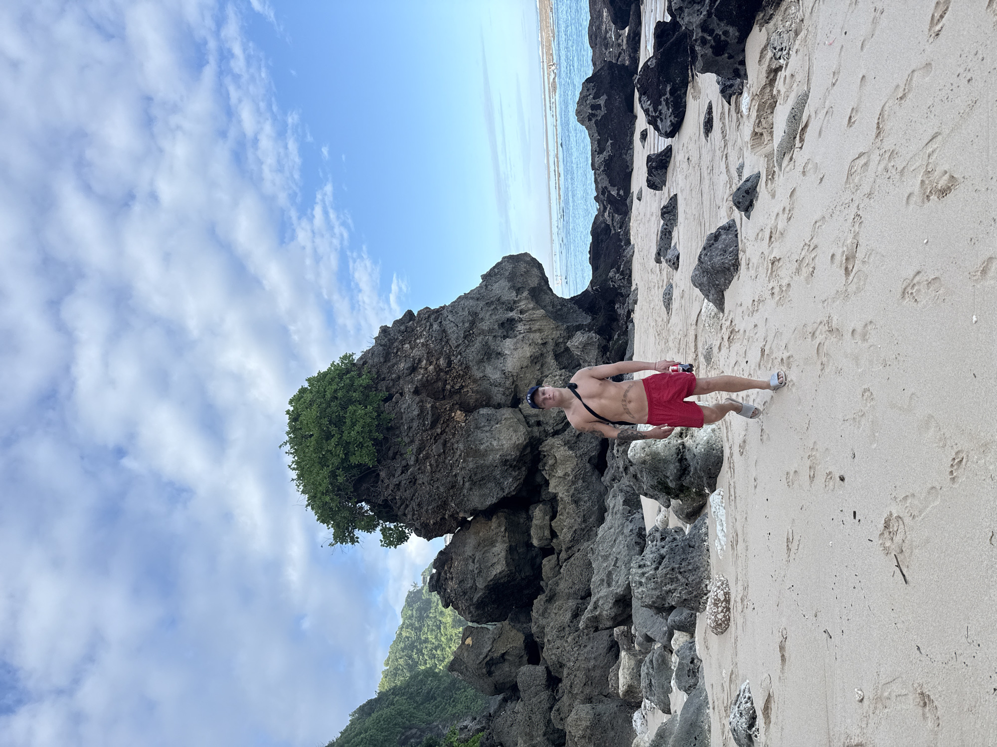 The image depicts a man standing on a sandy beach, likely in Kuta Selatan, Indonesia. He is wearing red shorts, a dark-colored baseball cap, and sandals. He is shirtless, revealing tattoos on his torso. A black strap crosses his chest and shoulder, likely attached to a bag or camera. He is standing near a large rocky outcropping, partially covered with vegetation. Beyond the rocks, the sea can be seen meeting the horizon under a partially cloudy sky. The beach itself is marked with footprints.