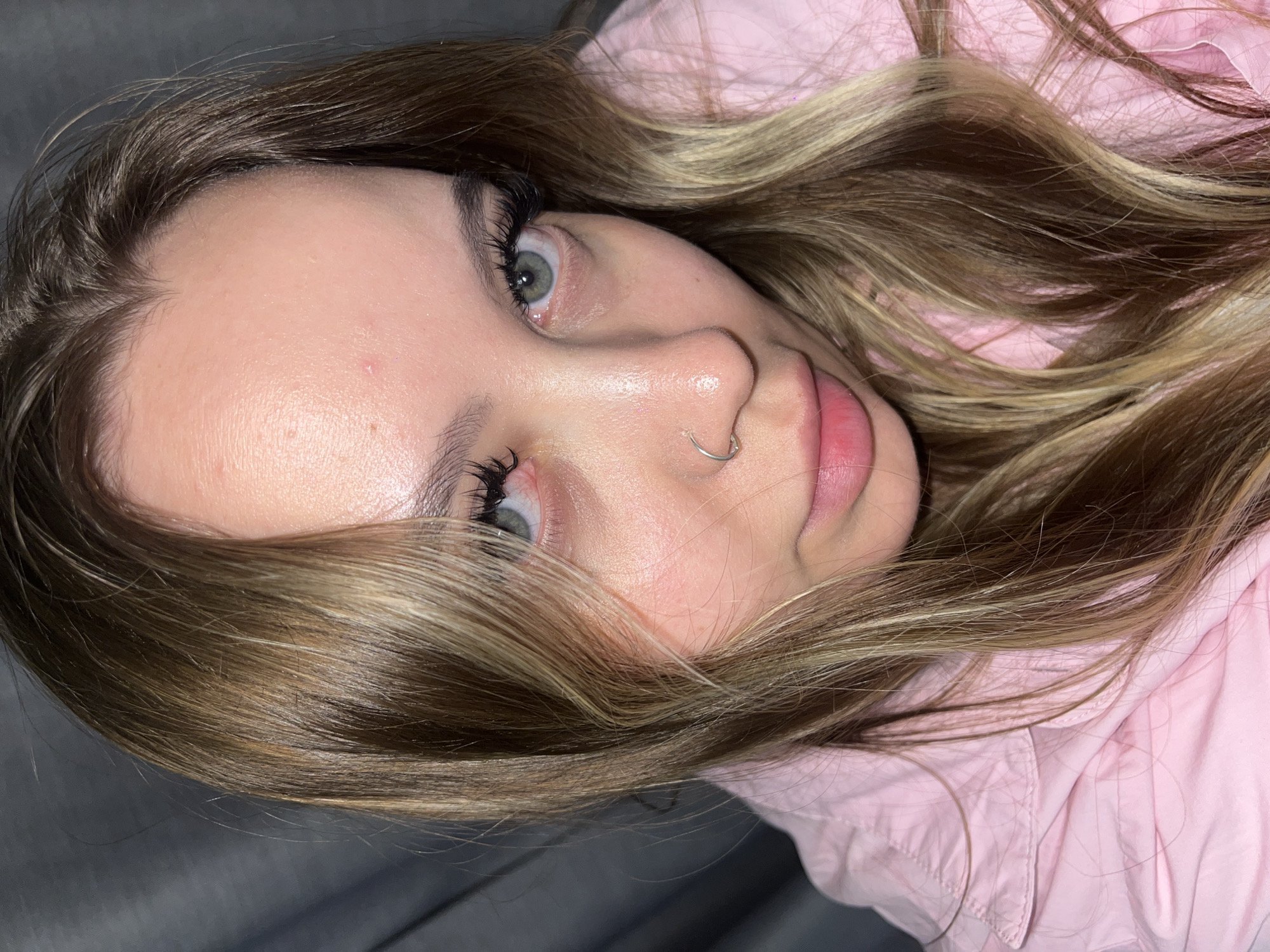 A young woman lies on a gray surface, perhaps a couch or bed, wearing a light pink shirt or jacket. She has long brown hair with blonde highlights. Her eyes are light, possibly green or grey. She is wearing makeup, specifically long eyelashes, and a nose ring. The image provides no information about the city of Krasnodar or Russia.