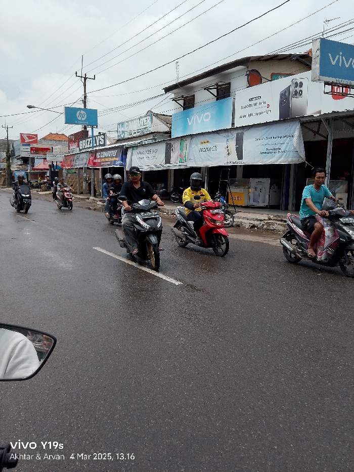 The image shows a street in Jatibarang, Indonesia, with several people riding motorcycles. The street is wet, likely from recent rain, and there are several shops along the street. The shops sell a variety of goods, including electronics and computers. The shops have signs advertising Vivo phones and HP computers. The motorcycles are all traveling in the same direction, and the riders are wearing helmets. The image was taken on March 4, 2025, at 1:16 PM. The image shows the street is busy with traffic, and the riders are traveling at a moderate pace. The buildings along the street are fairly close together. The buildings appear to be in good condition, and the street is relatively clean.