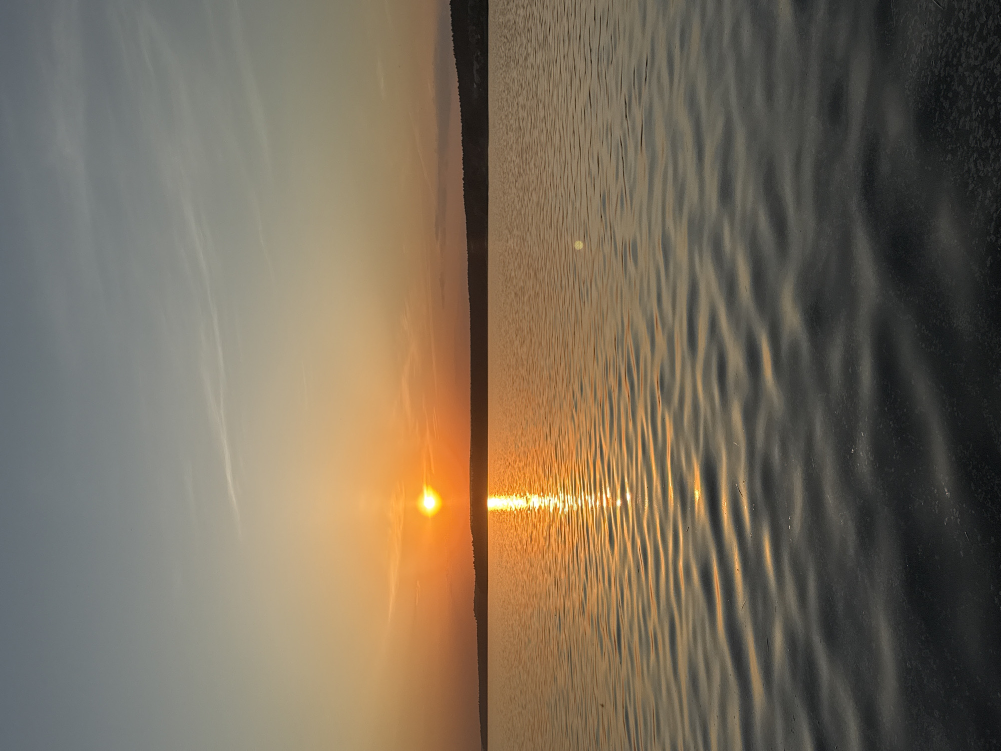 The image captures a sunset scene, likely over a body of water. The left side of the frame depicts a gradient sky transitioning from a light blue to a warm orange, with thin wisps of clouds. In the middle, a bright sun is setting near the horizon, casting an intense orange glow. A dark, seemingly forested landmass stretches along the horizon line. To the right, the water reflects the golden light of the sunset, creating rippling patterns and highlights. The water appears calm with subtle textures caused by the interplay of light and wavelets. The image suggests a peaceful, natural setting. While the location is specified as Votkinsk, Russia, without further information, I cannot confirm if the water is from the Votkinsk reservoir or another nearby body of water. There are no people or objects visible in the frame besides the natural elements.