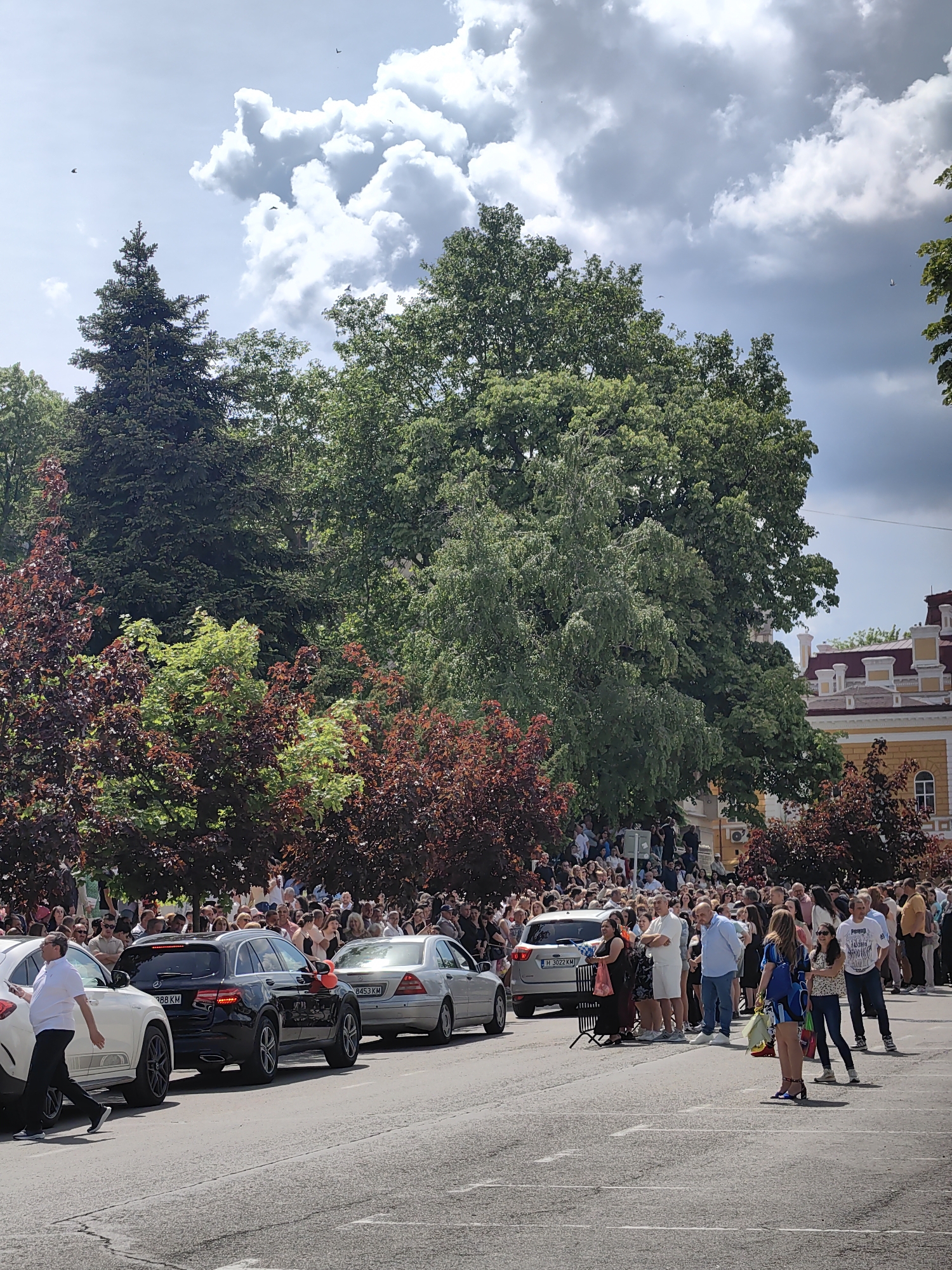 The image captures a street scene in Shumen, Bulgaria, likely during an event or gathering. A large crowd of people stands along the street. On the left side of the street, several cars are parked bumper to bumper, including a white SUV, a black Mercedes SUV, and a silver sedan. The black Mercedes has a red balloon attached to it. Many people are standing on the sidewalk or at the edge of the street, some of whom are looking at the parked cars. There is a black metal barricade to the right of the silver sedan.
The background features lush greenery, including tall trees and bushes with red foliage. Above the trees is a partially cloudy sky. The architecture of a building with a yellow facade and red-tiled roof is visible on the right side of the image. The overall scene indicates a public event of some kind, possibly a celebration or gathering that has drawn a significant crowd.