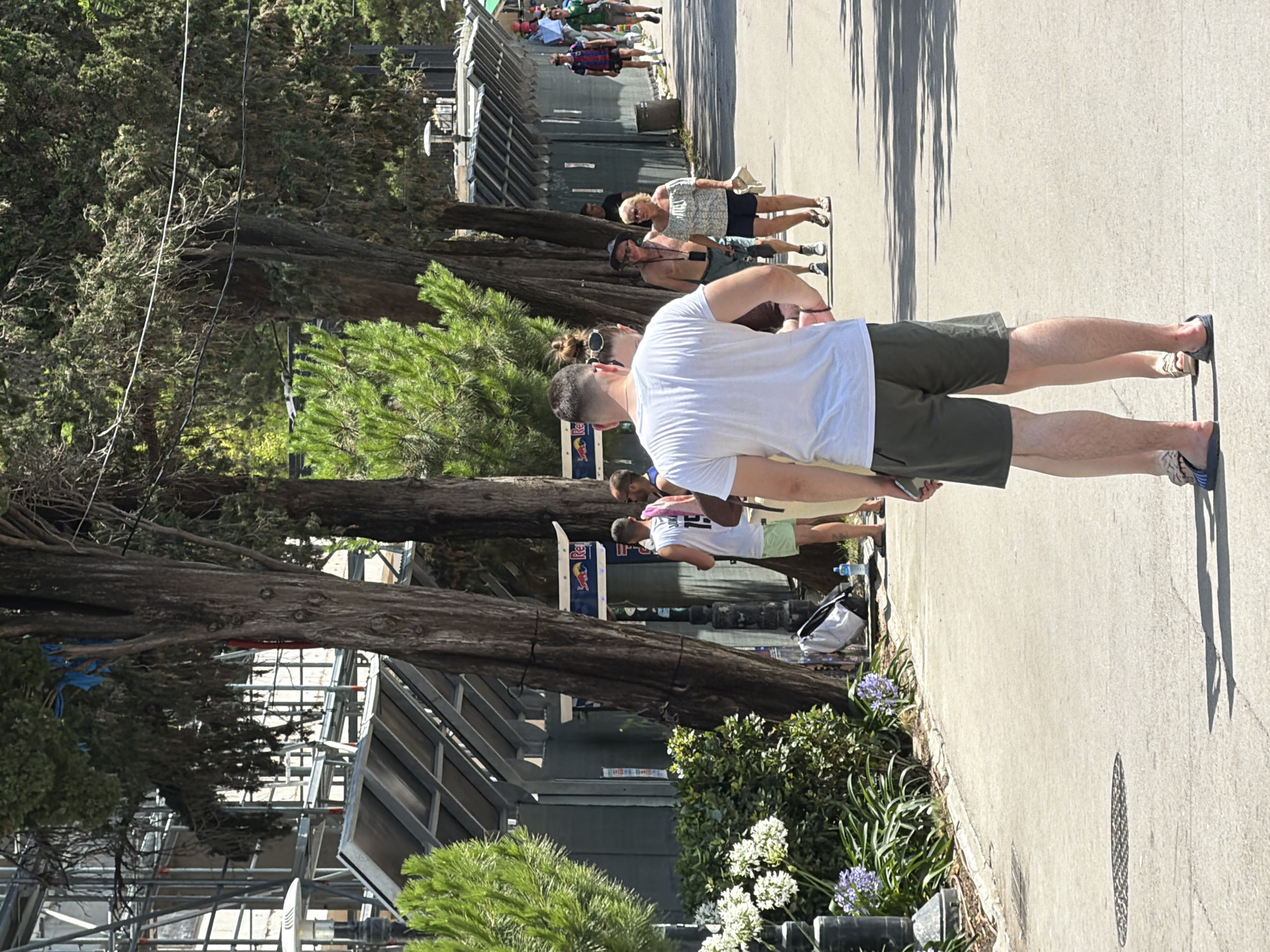 In Budva, Montenegro, a paved walkway is lined with tall, mature trees casting dappled shadows. The walkway hosts several people walking in various directions. In the foreground, a man in a white t-shirt and dark shorts stands facing away from the camera, with his back to the viewer. To his left and slightly behind him, a woman in a similar outfit is partially visible. Further down the walkway, other individuals are seen walking, including a man with a blue towel over his shoulder and a woman in a white off-the-shoulder top and dark shorts. In the background, a row of structures with metallic roofs are visible, some adorned with Red Bull signage. The environment appears to be an outdoor recreational area or a public thoroughfare within the city.