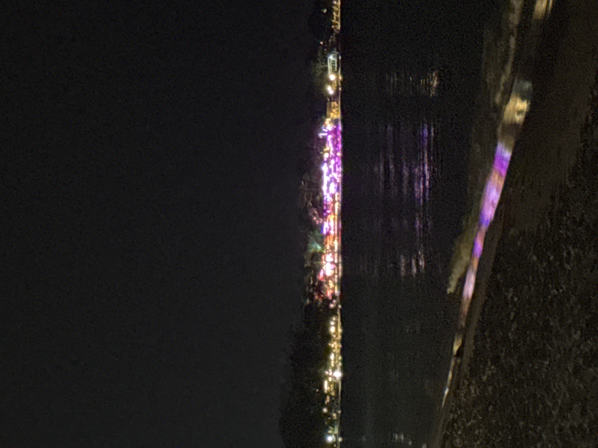 This media file shows a nighttime scene in Ko Samui, Thailand, likely along a waterfront or street with buildings decorated with lights. The dominant colors are the deep black of the night sky and water, contrasted with the vibrant, multi-colored lights. These lights, primarily in shades of purple, pink, yellow, and white, illuminate a narrow strip of what appears to be a building or a festive display.
The lights are arranged vertically, suggesting they are part of a structure like a tower, a series of connected buildings, or possibly even a large art installation. Some of the lights reflect on the dark surface of the water, creating shimmering patterns that add to the festive and dynamic atmosphere. The surface of the water appears calm, with the reflections being distinct.
On the right side of the image, there is a textured, dark surface that could be a wall, a rocky embankment, or the side of a building. A thin, vertical band of colorful lights, similar to those seen on the left, is also visible on this surface, indicating the decorations extend across different elements of the scene.
The scene evokes a sense of celebration or a special event, possibly a festival or a holiday, given the abundance and variety of decorative lights. The time of day is clearly night, with no natural light visible. The location cues, such as "Ko Samui District, Thailand," suggest a tropical setting, though specific environmental details like weather are not evident beyond the darkness and the presence of water. No people are clearly identifiable, but the lights suggest human activity and a populated area. There is no visible text in the image.