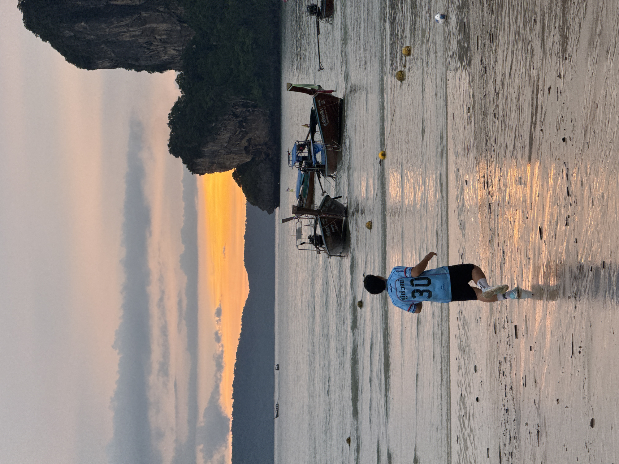 In Ban Ao Nang, Thailand, a person is captured mid-stride running on a sandy beach during twilight. The individual wears a light blue jersey with the number 30 prominently displayed in dark blue, paired with black shorts. The sun is setting, casting a warm orange glow across the sky, which is partially obscured by scattered clouds. The sea is calm, with several longtail boats anchored offshore. Yellow buoys are visible in the water, marking an area. The wet sand reflects the sky's colors, creating shimmering patterns. In the background, a large, verdant cliff face rises from the sea, characteristic of the region's karst landscape. The scene depicts a solitary moment of physical activity against a scenic, natural backdrop.