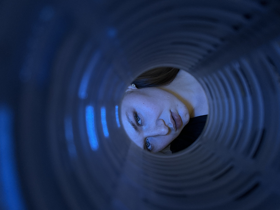 A woman's face peers through a circular opening, possibly a pipe or tunnel, viewed from a unique perspective. The scene is bathed in cool, blue lighting, accentuating the circular form surrounding the subject. The concentric rings lead to the woman's face, featuring light skin, subtle makeup, and visible facial features. The image conveys a sense of confinement or observation, with the woman's gaze directed toward the viewer.