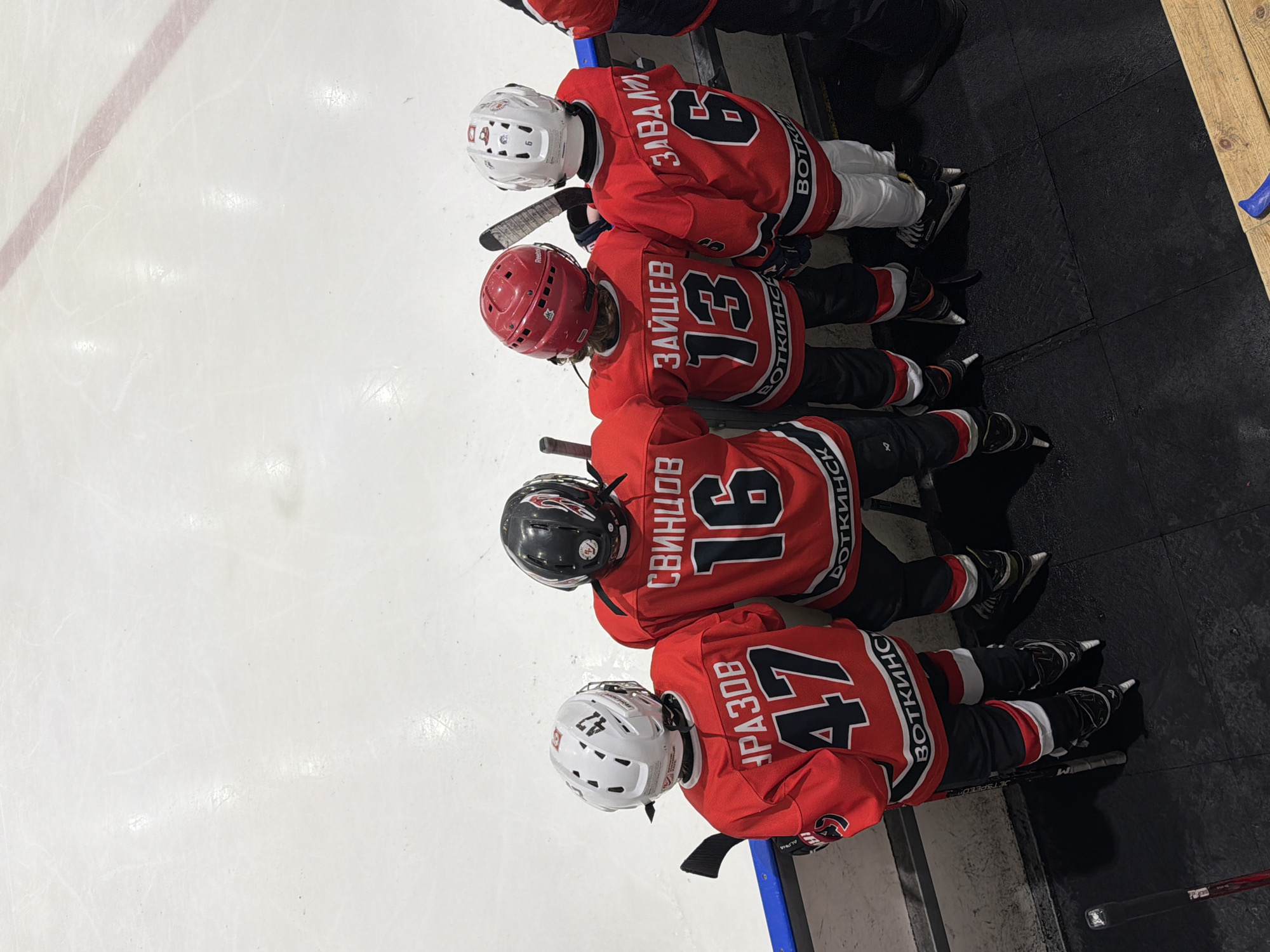 The image shows an overhead view of four young ice hockey players, wearing red, black, and white uniforms, seated on a bench or barrier alongside an ice rink. They are positioned with their backs to the camera, looking out towards the ice.
**Subjects:** Four young hockey players are the main subjects. They are equipped with hockey helmets (white, red, and black), jerseys, and skates. Hockey sticks are visible near some of the players.
**Activity or event:** The players appear to be resting or waiting on the team bench during a hockey practice, game, or event. Their posture suggests they are observing the ice.
**Notable details:**
* **Jerseys and numbers:** From right to left, the players' jerseys display the following names and numbers in Cyrillic script:
* Player 1 (partially visible): Number 9, name "ЗАВАЛЕ" (Zavale).
* Player 2: Number 13, name "ЗАЙЦЕВ" (Zaytsev).
* Player 3: Number 16, name "СВИНЦОВ" (Svintsov).
* Player 4: Number 47, name "УРАЗОВ" (Urazov). This player's white helmet also has "47" prominently displayed.
* **Team Name:** All visible jerseys have "ВОТКИНСК" (Votkinsk) printed vertically along the side, indicating their team's origin or name.
* **Setting:** The left side of the frame shows the smooth, white ice surface, marked with skate lines and a red line. The right side features a darker area with black protective mats and wooden planks, characteristic of a player bench or penalty box area in an ice arena.
* **Time/Weather:** The indoor setting and artificial lighting indicate no specific time of day or weather conditions.
* **Emotions:** The players' faces are not visible, so emotions cannot be determined.
The scene depicts a typical moment in youth ice hockey, with players waiting for their turn on the ice or taking a break from play. The text "ВОТКИНСК" confirms the location context of Votkinsk, Russia.