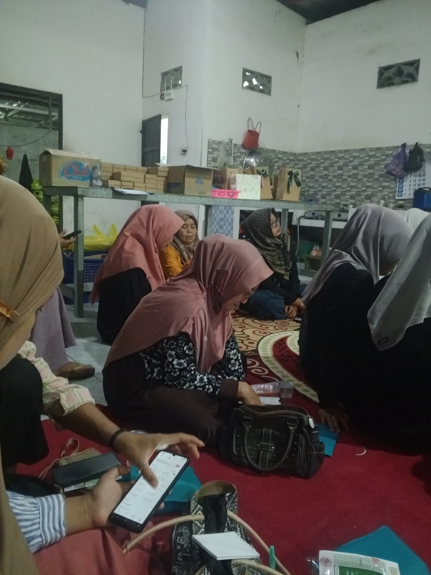 The image shows a gathering of at least five women sitting on a red carpeted floor indoors, in what appears to be a residential or community space in Mlilir, Indonesia. The room has white walls and a tiled backsplash in the background, suggesting a kitchen or communal area. Some of the women are wearing hijabs. In the foreground, one woman is holding and looking at a smartphone. Several bags, including a dark brown handbag, are placed on the floor near the women. In the background, there are shelves with boxes and other items, and a calendar is visible on the wall. The lighting is dim, indicating it could be evening or an indoor space with limited natural light.