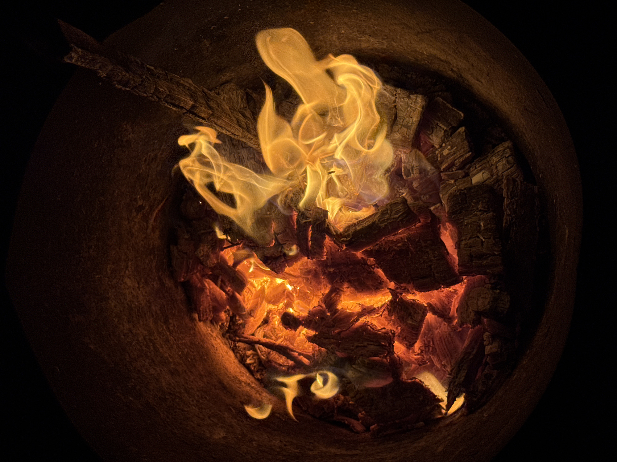 The image shows an up-close view of a fire burning in a fire pit, likely located in the city of Chaikovsky, Russia. The fire is contained within a roughly cylindrical structure, possibly made of metal or stone, with a rough, textured interior. The pit is filled with glowing embers and burning logs, producing vibrant orange and yellow flames that twist and flicker. One log, partially consumed by the fire, juts out towards the right side of the frame. The background is dark, suggesting it is nighttime or the interior of the pit is in shadow. The overall impression is one of intense heat and dynamic movement of the flames.