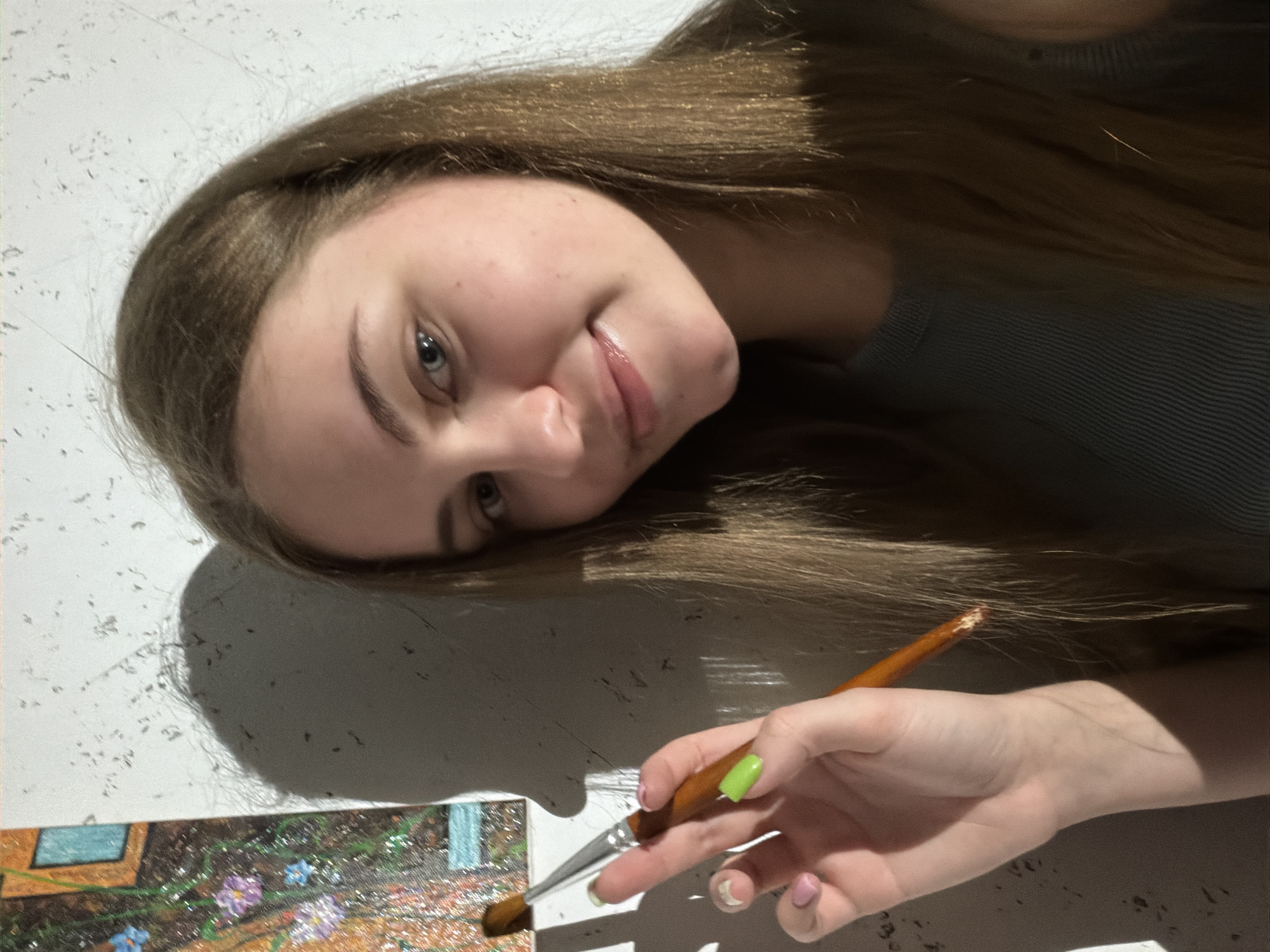 A young woman with long, brown hair and blue eyes is holding a paintbrush with a brown tip in her right hand. She is wearing a gray, ribbed top. Her fingernails are painted in varying shades of pink and green. She is standing in front of a textured wall, with a shadow cast from her hand and an unseen object. To her left, a painting with floral elements and a window can be seen. The scene appears to be indoors, possibly an art studio or a room in a residence. The background wall has a white, textured surface, and the lighting suggests an indoor setting. The overall composition is a close-up portrait. No specific location in Dnipro, Ukraine, can be determined from the provided image.