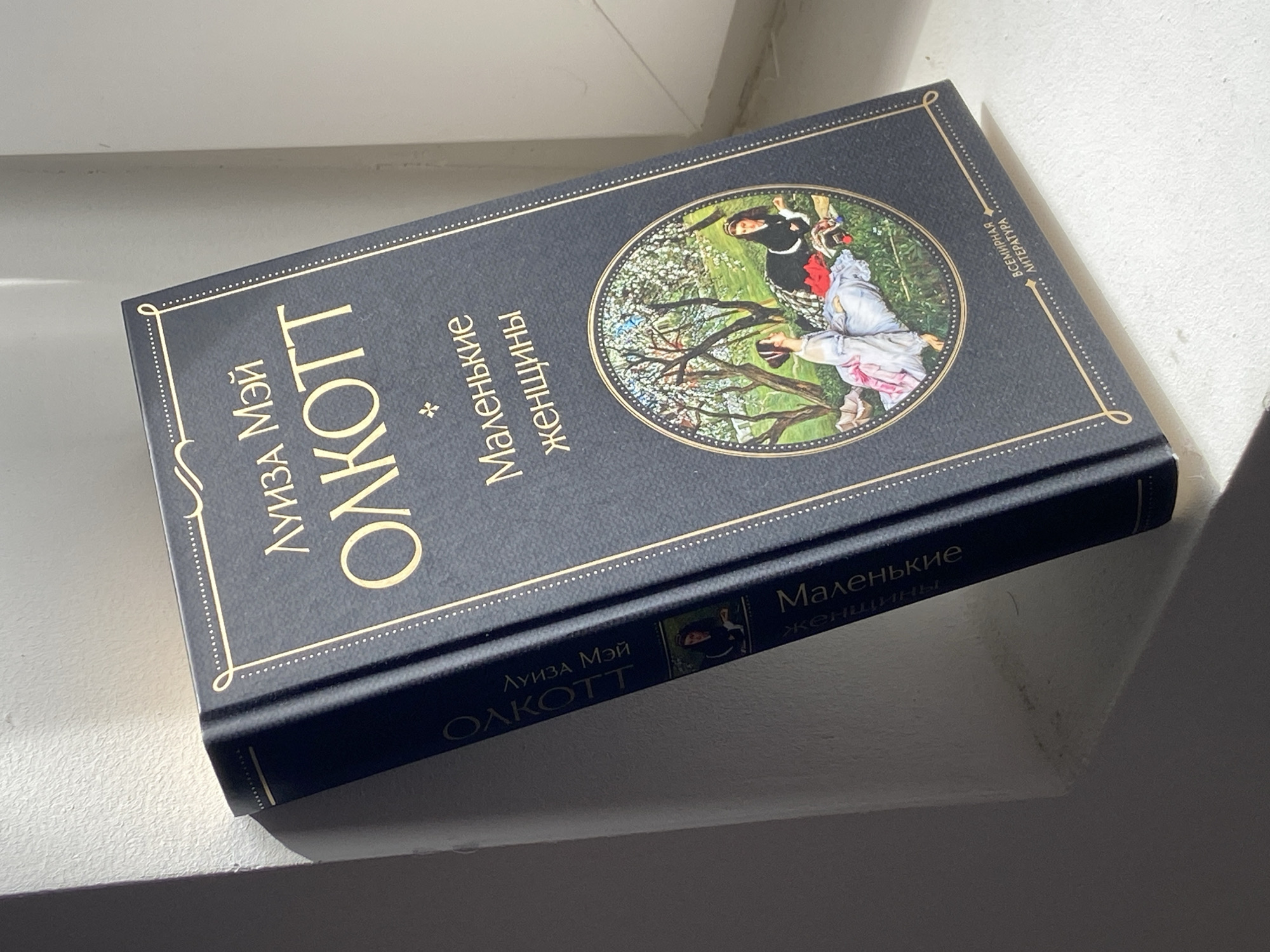 A dark blue hardcover book, identified as a Russian edition of Louisa May Alcott's "Little Women," lies diagonally on a light-colored, textured surface, possibly a windowsill or shelf. Strong sunlight from an unseen window casts distinct shadows to the right of the book, indicating it is daytime.
The book's cover is decorated with gold foil lettering and intricate borders. A central circular illustration depicts two young women and a man in a vibrant outdoor setting with green grass and blossoming trees, suggestive of spring or summer. The figures are dressed in period clothing; one woman is shown reaching out while another looks upwards, with the man standing nearby.
The author's name, "ЛУИЗА МЭЙ ОЛКОТТ" (Louisa May Alcott), and the title, "Маленькие женщины" (Little Women), are clearly visible in gold on the front cover. The spine also features the author's name and title, along with a small inset image derived from the main cover illustration. Text on the upper right corner of the front cover reads "ВСЕМИРНАЯ ЛИТЕРАТУРА" (World Literature). The scene suggests a quiet, sunlit indoor setting where Russian is spoken, consistent with the provided location context of Tyumen, Russia.