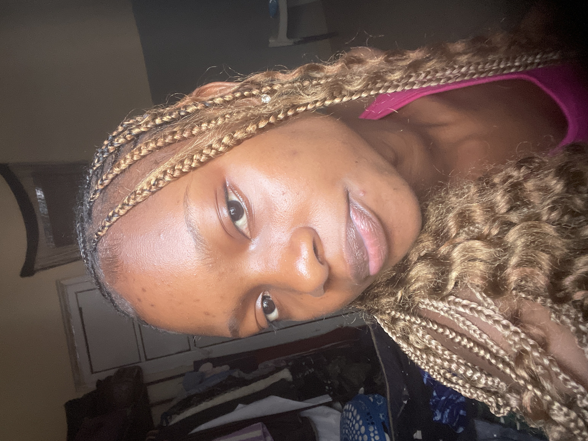 The image is a close-up, selfie-style photograph of a young Black woman with braided hair, taken in what appears to be an indoor, domestic setting. The woman is positioned centrally, with her face tilted slightly to her left. She has dark skin, prominent cheekbones, and her lips are slightly parted. Her hair is styled in numerous thin braids that are a mixture of dark brown and lighter blonde strands, with some braids adorned with small, sparkling accessories. She is wearing a bright pink top.
The background of the image is somewhat out of focus, but reveals elements of a room. To the left, there is a dark closet or wardrobe with clothes hanging inside. In the lower left corner, a blue and white patterned object, possibly a basket, is visible. The wall behind her is a muted color. Further to the right, a white chair can be seen. The lighting suggests it is daytime, with light sources illuminating the subject's face, creating a subtle sheen on her skin.
The provided hints show close-ups of the woman's face, hair, and clothing, as well as parts of the background environment, such as the closet and a door.
The prompt requests the location to be specified as Lafia, Nigeria. Without any contextual information within the image itself, it is impossible to definitively confirm the location. Therefore, the following description adheres to the prompt's request to include this information as stated, while acknowledging the lack of visual evidence to support it.
The scene is located in Lafia, Nigeria. The photograph captures a young woman in her home, possibly a bedroom or living area, given the presence of personal belongings and furnishings. The lighting and composition suggest a casual, personal moment captured by the subject herself.