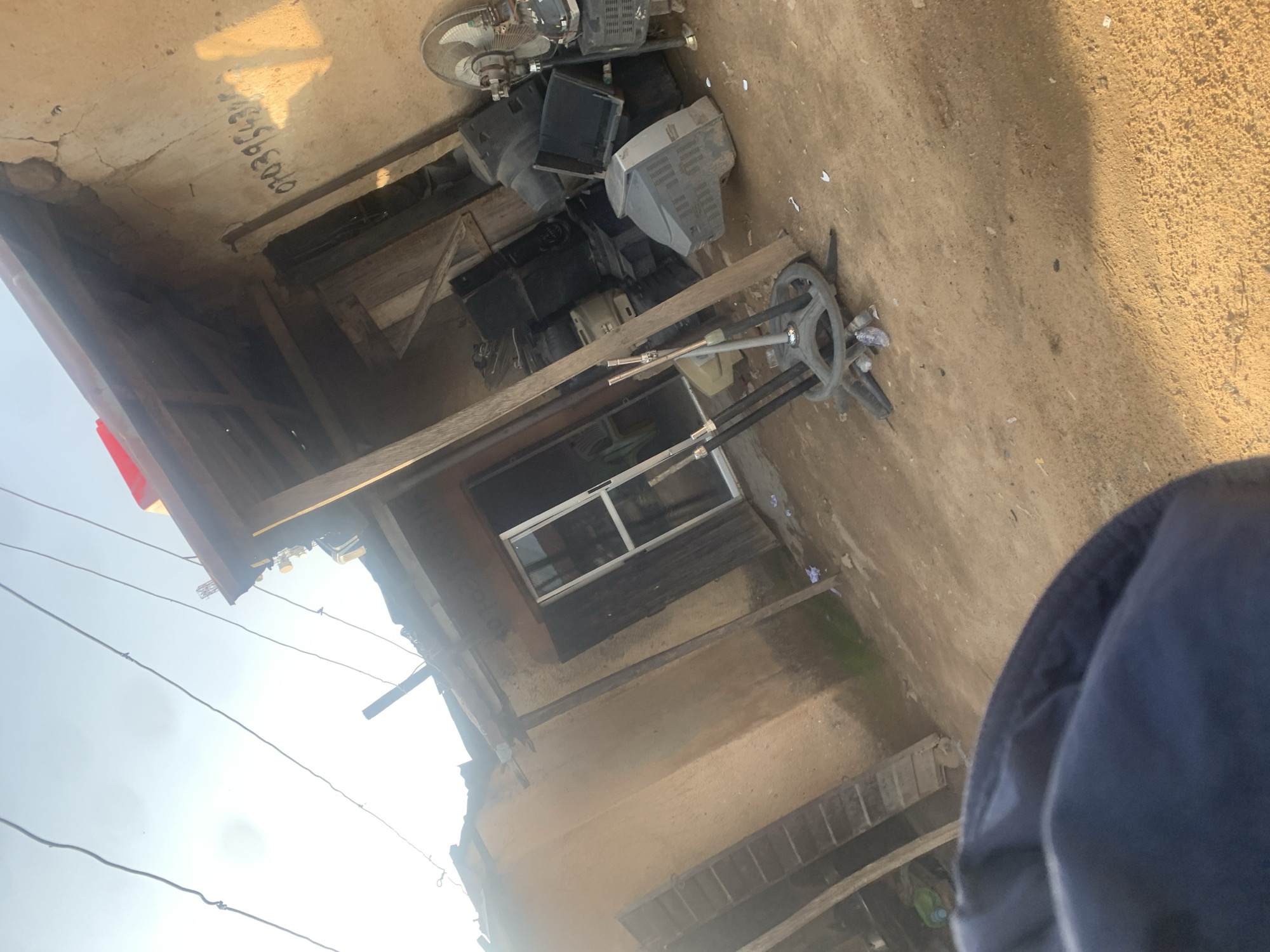 The image depicts an outdoor scene in Jos, Nigeria, featuring a collection of discarded electronic items, primarily old television sets and a fan, piled on the ground near a rustic structure. The setting appears to be an informal area, possibly a junkyard or a storage space for scrap materials. The ground is dusty and uneven, with a light-colored concrete slab visible in the upper portion of the image, possibly part of a building or walkway.
The electronic items are stacked haphazardly, with some television sets showing signs of wear and tear. A fan is positioned at the top left, and a circular base with protruding metal rods, possibly from a fan stand or other equipment, is visible in the foreground. A window with a metal frame is set into the wall of the structure. The overall impression is one of disarray and neglect.
The lighting suggests it is daytime, with sunlight casting shadows on the concrete surface. The sky is overcast or hazy, as indicated by the bright, diffused light. The presence of electrical wires strung overhead further points to a potentially developing or semi-developed area.
A visible detail on the concrete slab is a series of handwritten numbers in black ink, which read "07039553346". This appears to be a phone number.
The image is taken from a low angle, with a person's dark blue garment or jacket occupying the lower right corner, suggesting the photographer is in the scene. No distinct emotions are conveyed, but the scene itself evokes a sense of the mundane or the overlooked, typical of areas where discarded items are collected.