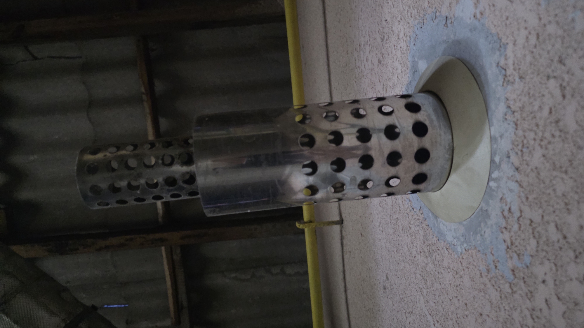 A close-up, low-angle shot depicts two perforated metal cylinders, likely parts of a ventilation or exhaust system, positioned vertically. The larger cylinder is at the bottom, resting on a beige, cone-shaped flange that is set into a rough, beige-colored surface. The smaller cylinder is stacked on top of the larger one. The background consists of a corrugated metal ceiling with wooden beams and a yellow pipe running horizontally across the middle ground. The scene appears to be indoors, possibly in a utility space or an unfinished building. The lighting is dim and directional, creating shadows and highlighting the metallic surfaces. No individuals are present in the image. The location in the city of Chuy, Kyrgyzstan, cannot be determined from the visual information provided.