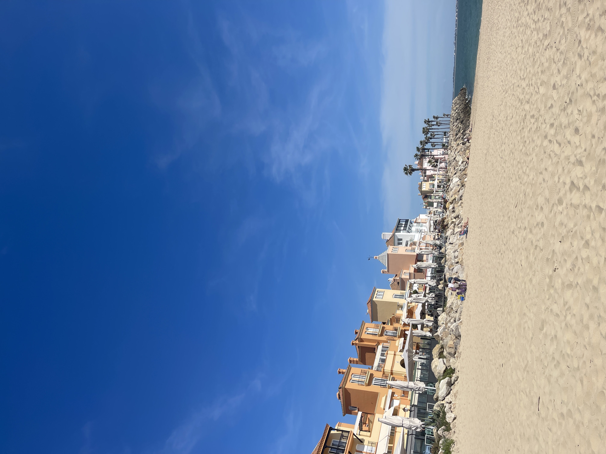 The image depicts a coastal scene in El Puerto de Santa María, Spain. On the right, a sandy beach occupies a significant portion of the frame. Adjacent to the beach is a series of buildings, mainly residential, constructed in a style typical of coastal Mediterranean architecture, featuring pastel colors and balconies. Palm trees are interspersed among the buildings. Between the buildings and the beach is a line of rocks, possibly serving as a breakwater or erosion control measure. In the background, there is a glimpse of the ocean horizon. The left side of the image is dominated by a clear blue sky with wispy clouds. The photo is taken from a low angle, enhancing the perception of height for both the buildings and the sky. The scene suggests a sunny day, with a calm atmosphere.