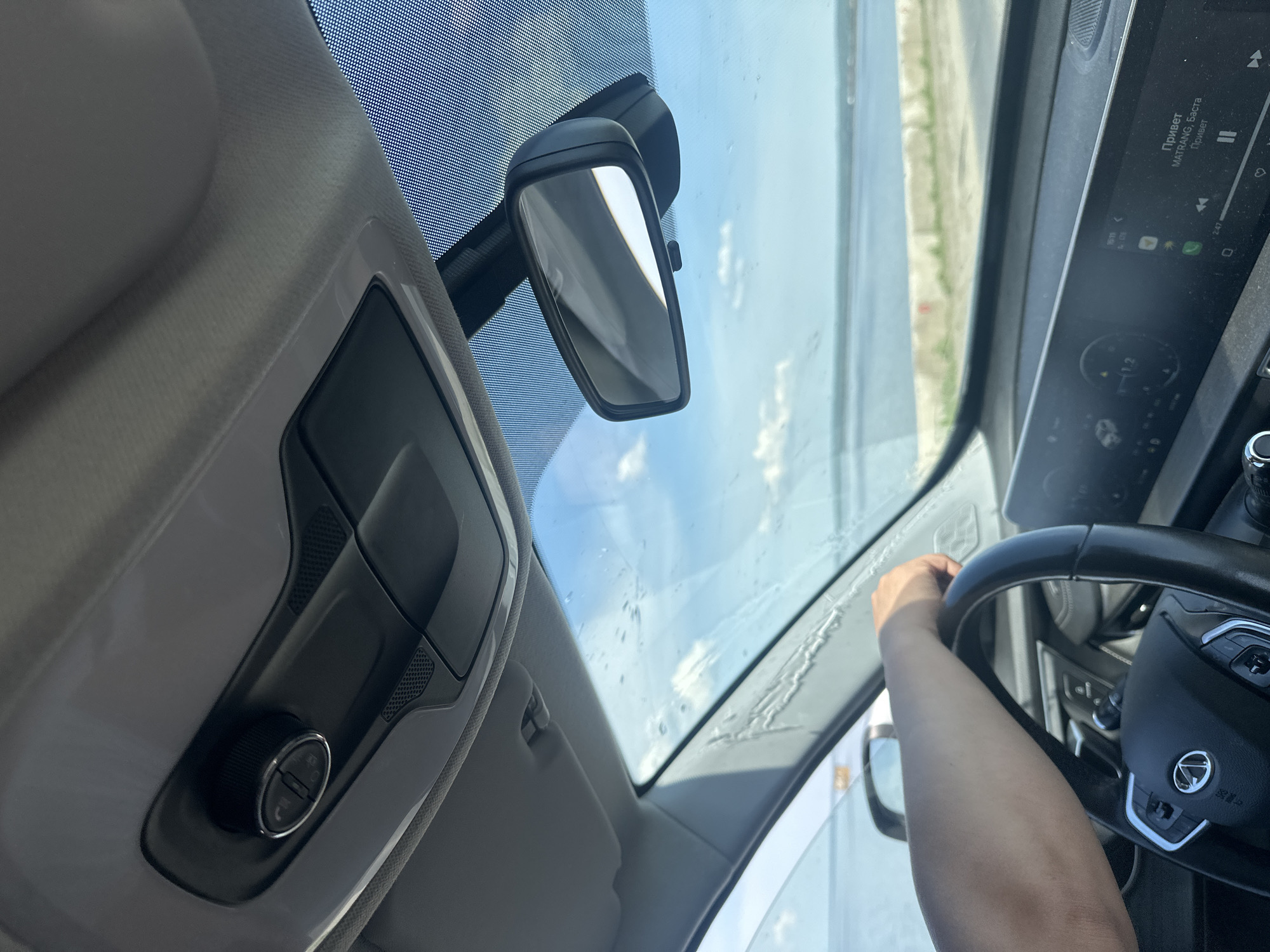 A view from inside a car, likely in motion. The driver's hand is visible on the steering wheel. The rear-view mirror is centrally located, with an overhead console to the left. The car's infotainment screen displays song information: "Привет" by MATRANG, Баста. The windshield shows a landscape with a light-colored ground, possibly sand or earth. The location is specified as Noviy, Russia, based on song titles being displayed in Russian.