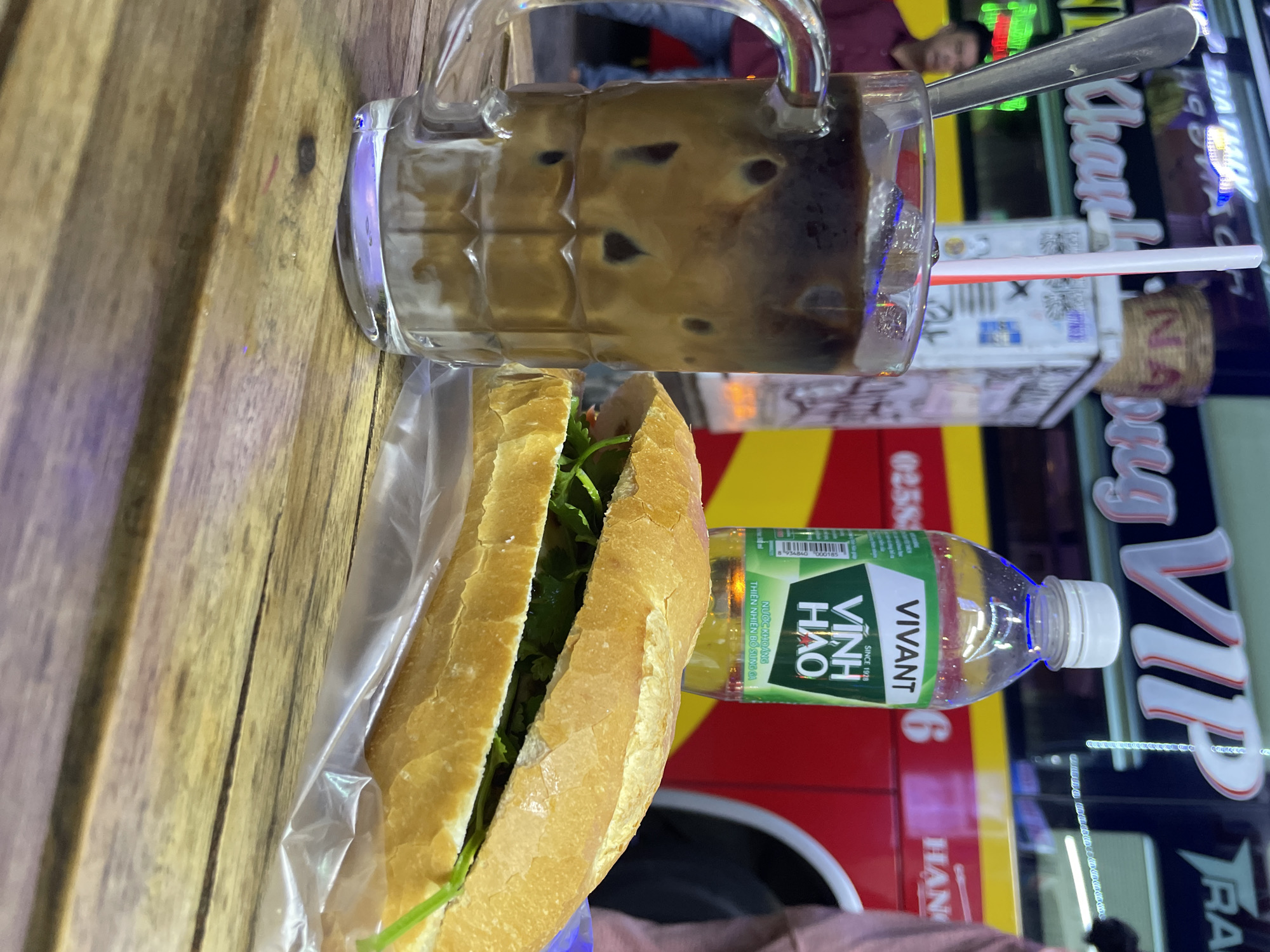 The image captures a meal consisting of a Vietnamese sandwich (banh mi), iced coffee, and a bottle of "VIVANT Vinh Hao" mineral water on a wooden table. The sandwich is wrapped in clear plastic. The coffee is served in a glass mug with a spoon and a straw. The mineral water bottle is mostly transparent. The background is blurred, but visible signs "ding VIP" and "HAN 6" suggest the image was taken in the city of Thủ Đức, Vietnam, and is a street scene with a bus or vehicle outside the frame. There is a person sitting behind the table.