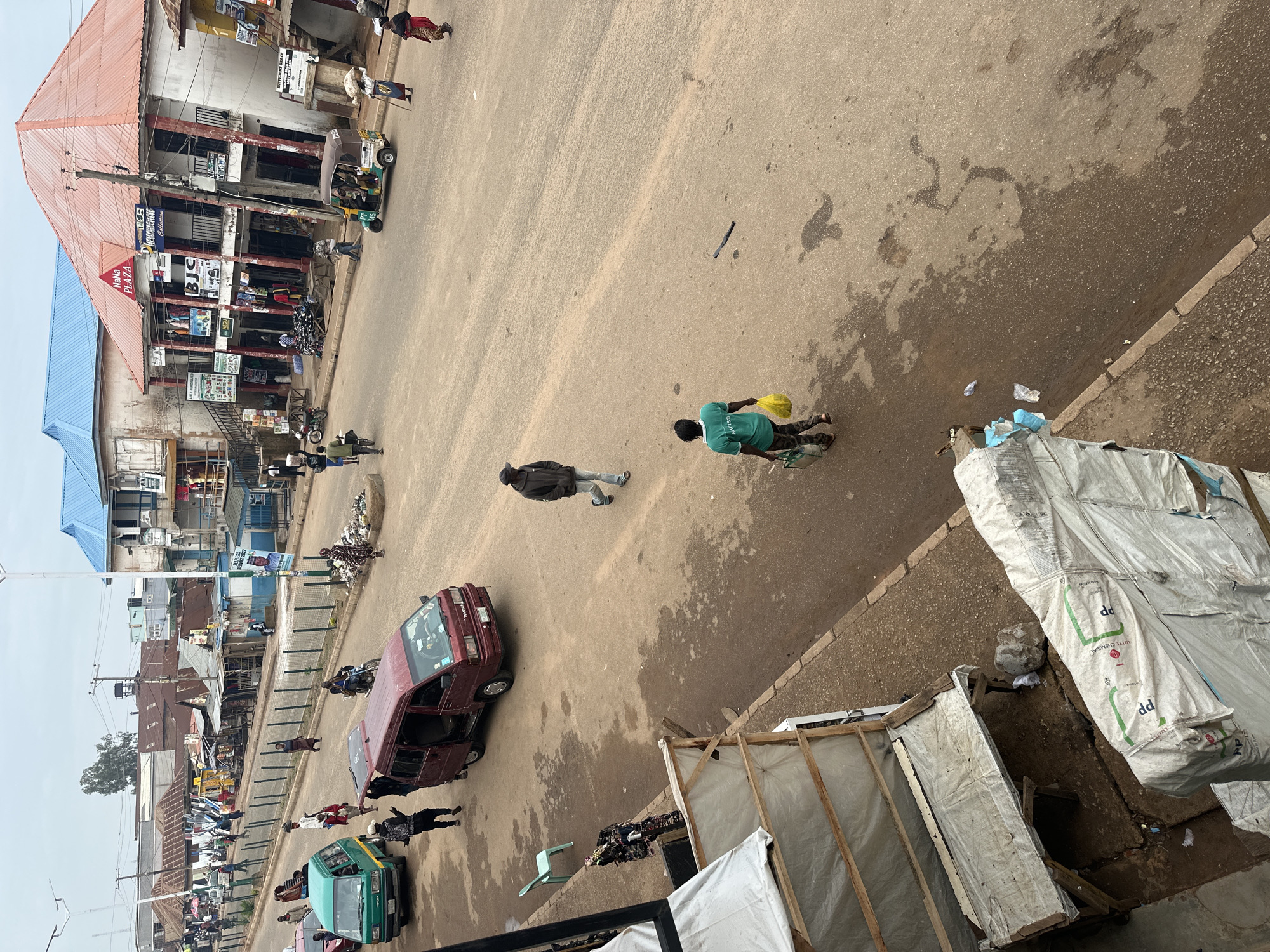 The image shows a street scene in Bukuru, Nigeria, under an overcast sky. A main paved road runs diagonally across the frame, with a smaller, rougher dirt path running alongside it. The road is populated by a variety of vehicles, including a green Toyota Hiace minibus and a burgundy Toyota Hiace minibus with its rear door open. A tricycle, known locally as a "Keke," is also present, carrying passengers. Several motorcycles are parked along the side of the road, and one person is riding a motorcycle.
Pedestrians are scattered throughout the scene, walking along the road and the dirt path. One man in a dark hooded jacket and baseball cap walks away from the camera on the road, wearing light-colored pants and sandals. Another person, wearing a bright green t-shirt and dark pants, carries two yellow shopping bags and walks towards the right side of the frame. A person in a patterned dress is seen carrying a bundle on their head.
Buildings line the sides of the road. On the right, a multi-story building with a red-tiled roof and a prominent sign reading "NaNa PLAZA" stands out, with various shops and businesses visible at street level. The buildings on the left appear to be a mix of single-story structures with corrugated metal roofs and some multi-story buildings. There are market stalls and open-fronted shops.
The ground is a mix of asphalt and dirt, with some areas appearing wet or muddy, suggesting recent rain. Debris, including plastic bags and what appear to be discarded items, is visible on the road and the edge of the sidewalk. Tarpaulins are used to cover some structures or goods. The overall impression is of a busy, lived-in urban environment.