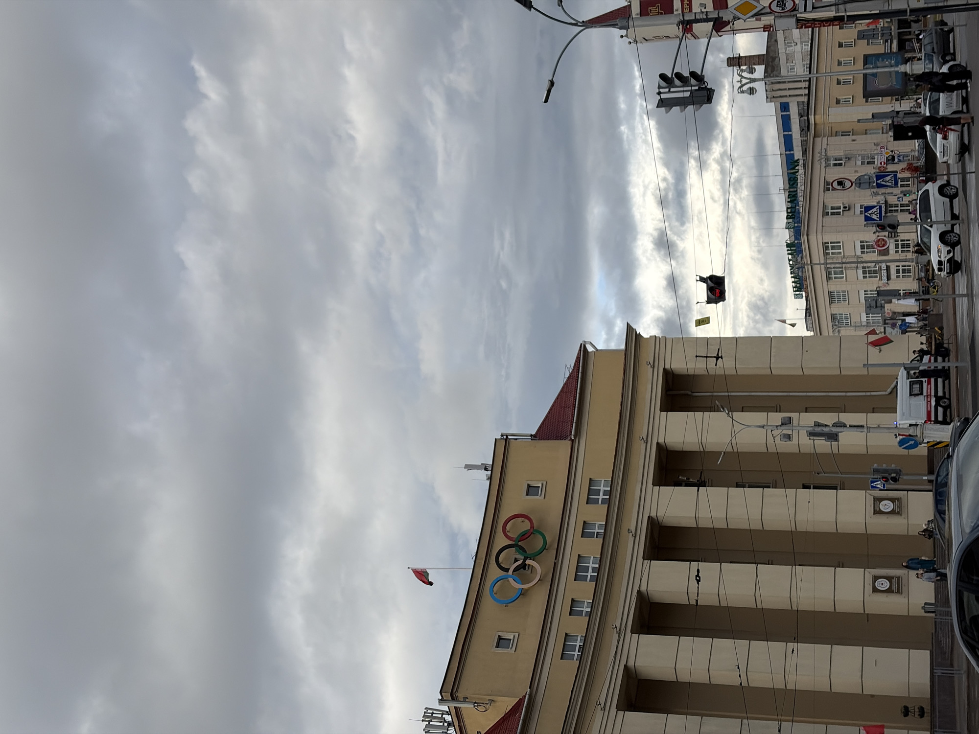 The image depicts an urban street scene in Minsk, Belarus, under a cloudy sky. Prominently featured is a large, pale yellow building with classical architectural elements, including tall columns. On the facade of this building, near the top, are the Olympic rings, colored blue, black, red, green, and yellow, along with a white flag bearing a red and white emblem flying from a flagpole.
In the background, other buildings are visible, including one with the Cyrillic text "БЕЛАРУСБАНК" (BELARUSBANK) on its facade. The street is busy with traffic, including several cars, one of which is a white van that appears to be an ambulance or emergency vehicle. Pedestrians are present on the sidewalks and crossing the street, and traffic lights are visible. The overall atmosphere is overcast and suggestive of an approaching or recent rain. The specific location is likely Independence Square (Ploshchad Nezavisimosti) in Minsk, given the presence of the Olympic rings on the former Government House and the BELARUSBANK building, which is adjacent to the square.