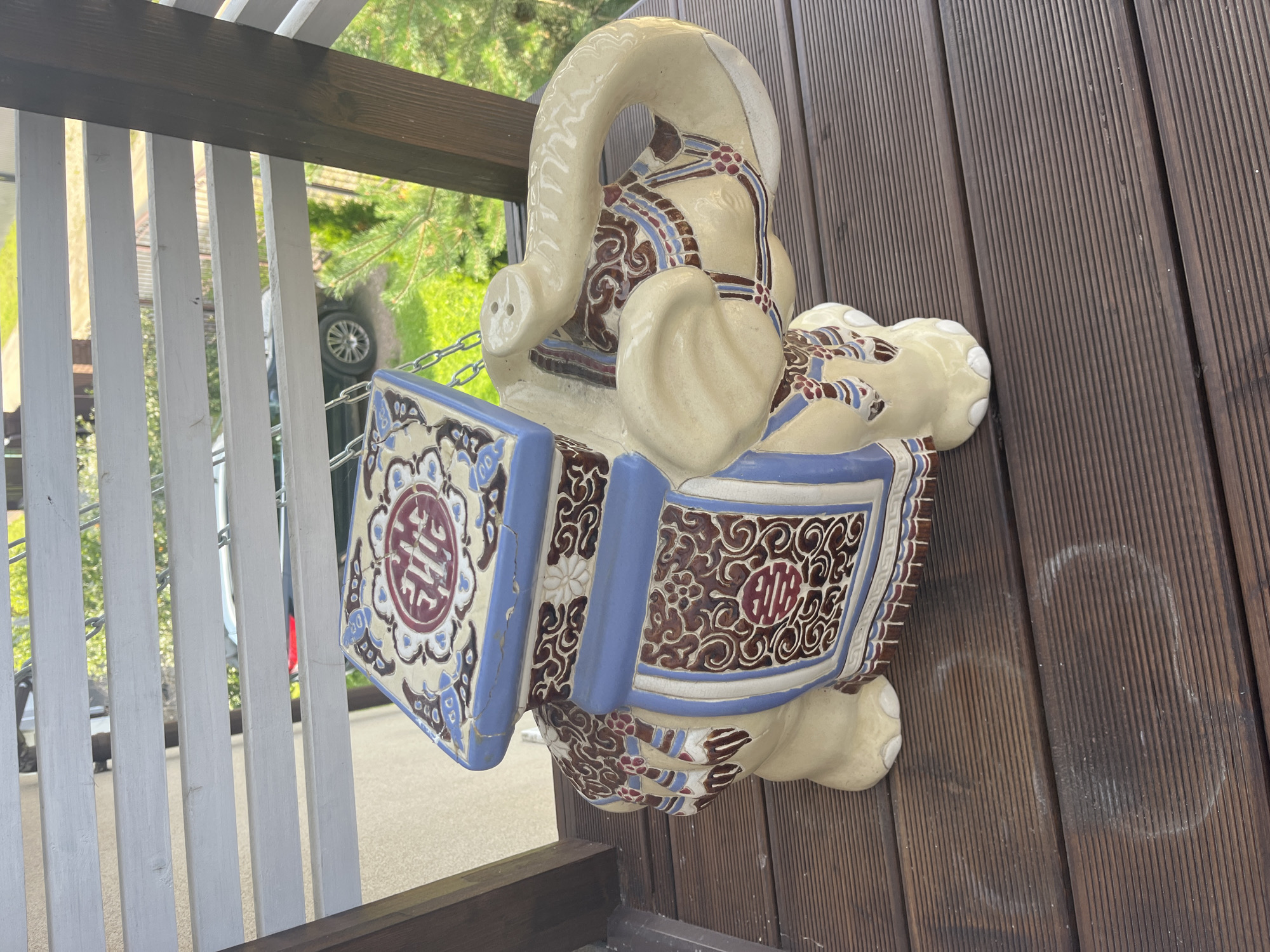 An ornate, ceramic elephant statue sits on a wooden deck. The statue is primarily cream-colored with blue, brown, and red decorative elements. The elephant has a saddle-like structure on its back, which is adorned with intricate patterns. The deck itself is made of dark brown wooden planks. In the background, through a slatted railing, a car is visible parked on a paved surface, and green foliage suggests an outdoor setting. The scene appears to be located on a balcony or patio.