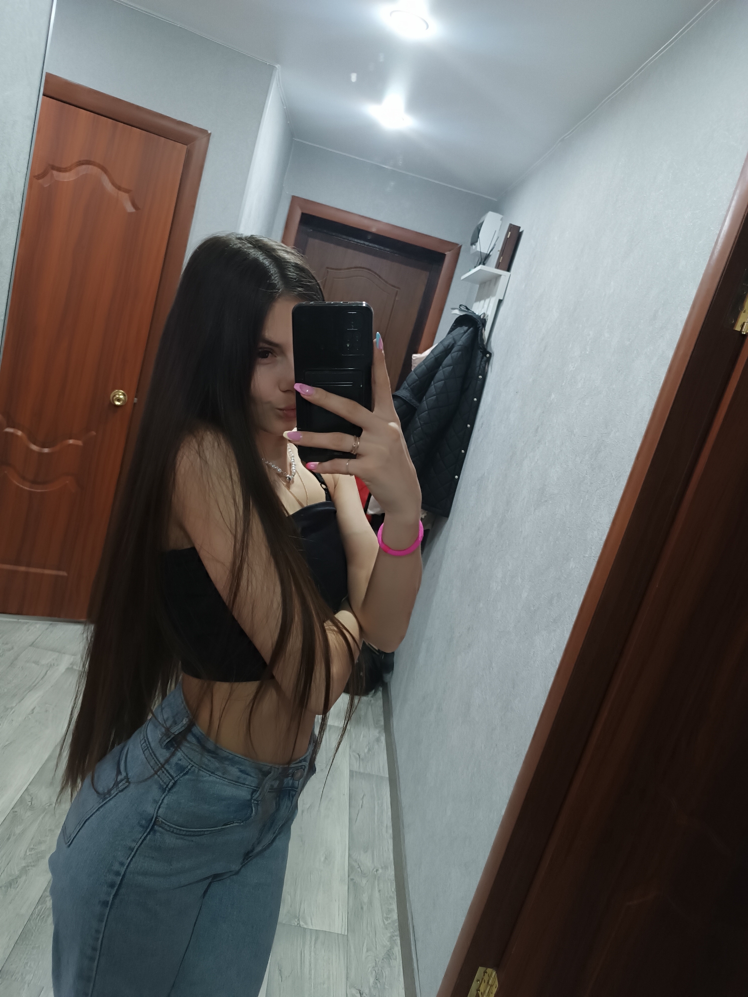 A woman is taking a selfie in a hallway. She is wearing a black top and light blue jeans. Her long, dark hair falls over her shoulder. She has a pink bracelet on her wrist. The hallway has wooden doors and light gray walls. A black jacket hangs on a shelf. The location is Unknown, Unknown.