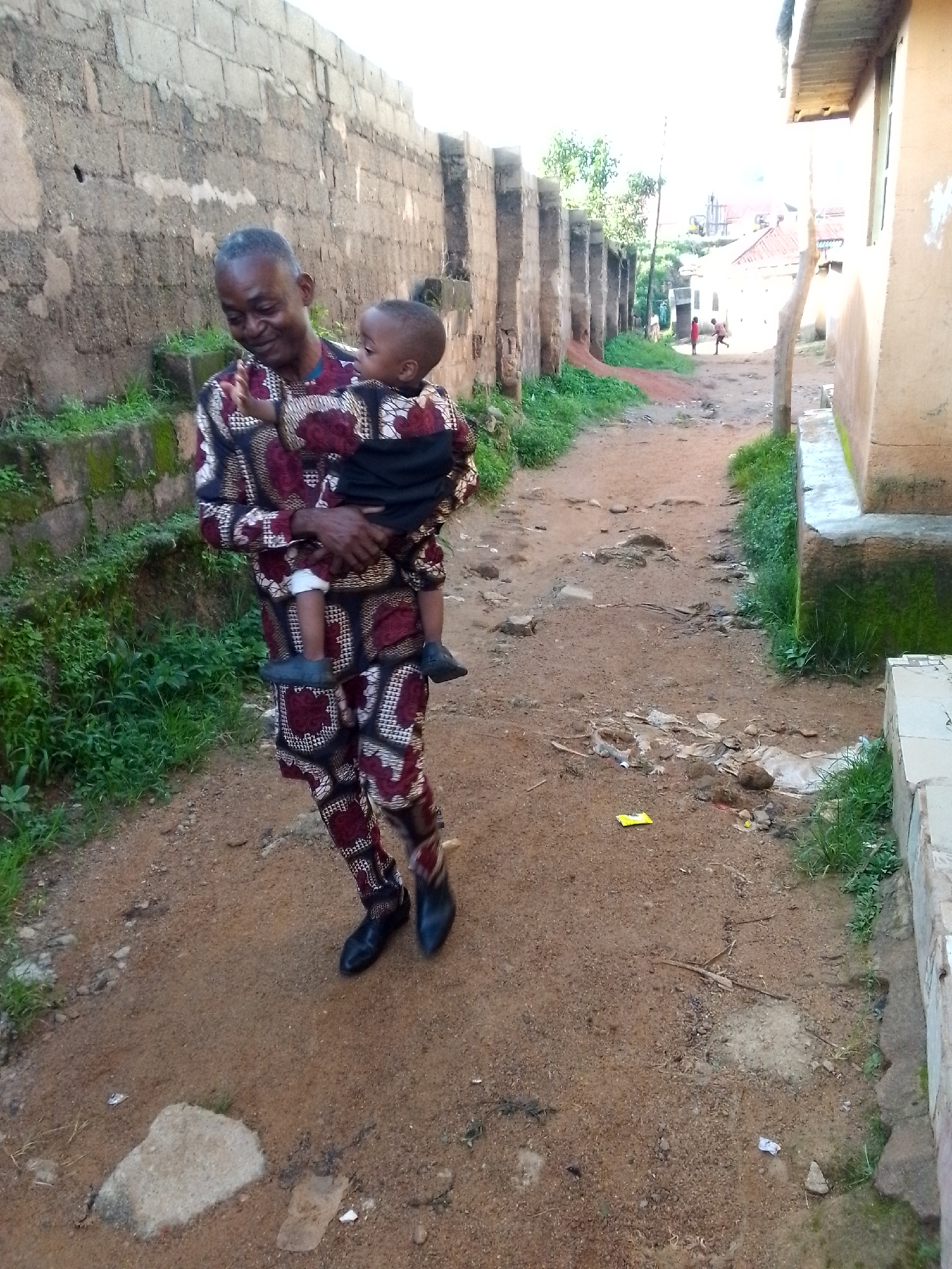 A middle-aged African man, wearing a patterned maroon and black dashiki and pants, carries a young African child, who is wearing a similar dashiki and black cloth. They are walking on a dirt path beside a moss-covered stone wall, which is part of a building. The path is uneven and littered with some debris, including a yellow packet and plastic. In the background, there are more buildings and trees, suggesting an urban or semi-urban environment in Jos, Nigeria. The man is looking down at the child with a gentle expression. The child is looking towards the left of the frame. The lighting suggests it is daytime, possibly late afternoon or early morning due to the softness of the light.