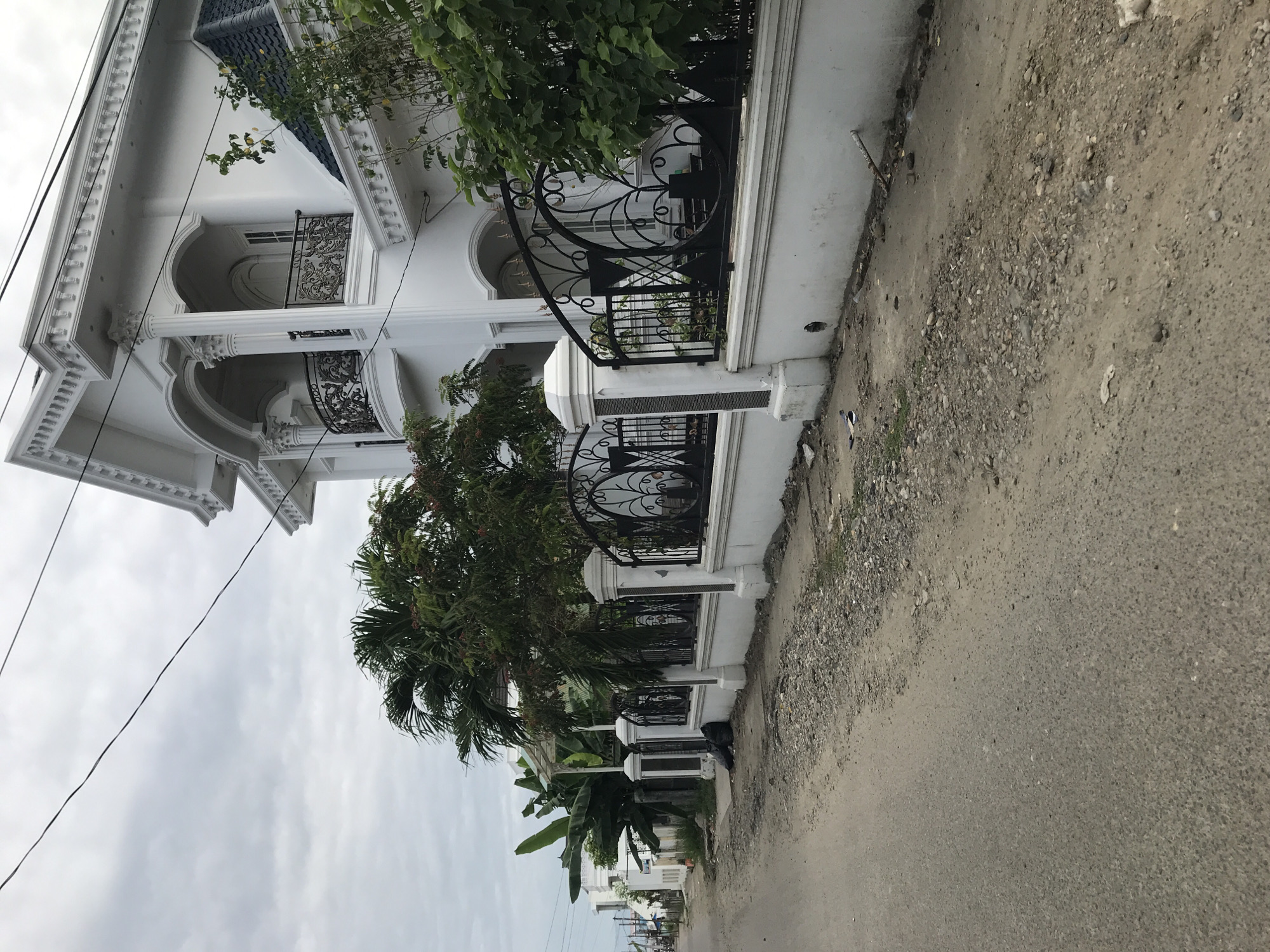 The image depicts a street view of a white, two-story building in Lhokseumawe, Indonesia. The building has elaborate architectural details, including arched balconies with ornamental black iron railings and decorative moldings. The exterior wall bordering the street is also white with black iron fencing. Lush greenery, including palm trees and leafy plants, obscures portions of the building and fence. A paved road borders the building. Overhead, electrical wires stretch across the sky. The sky is overcast. The scene appears to be a residential area.