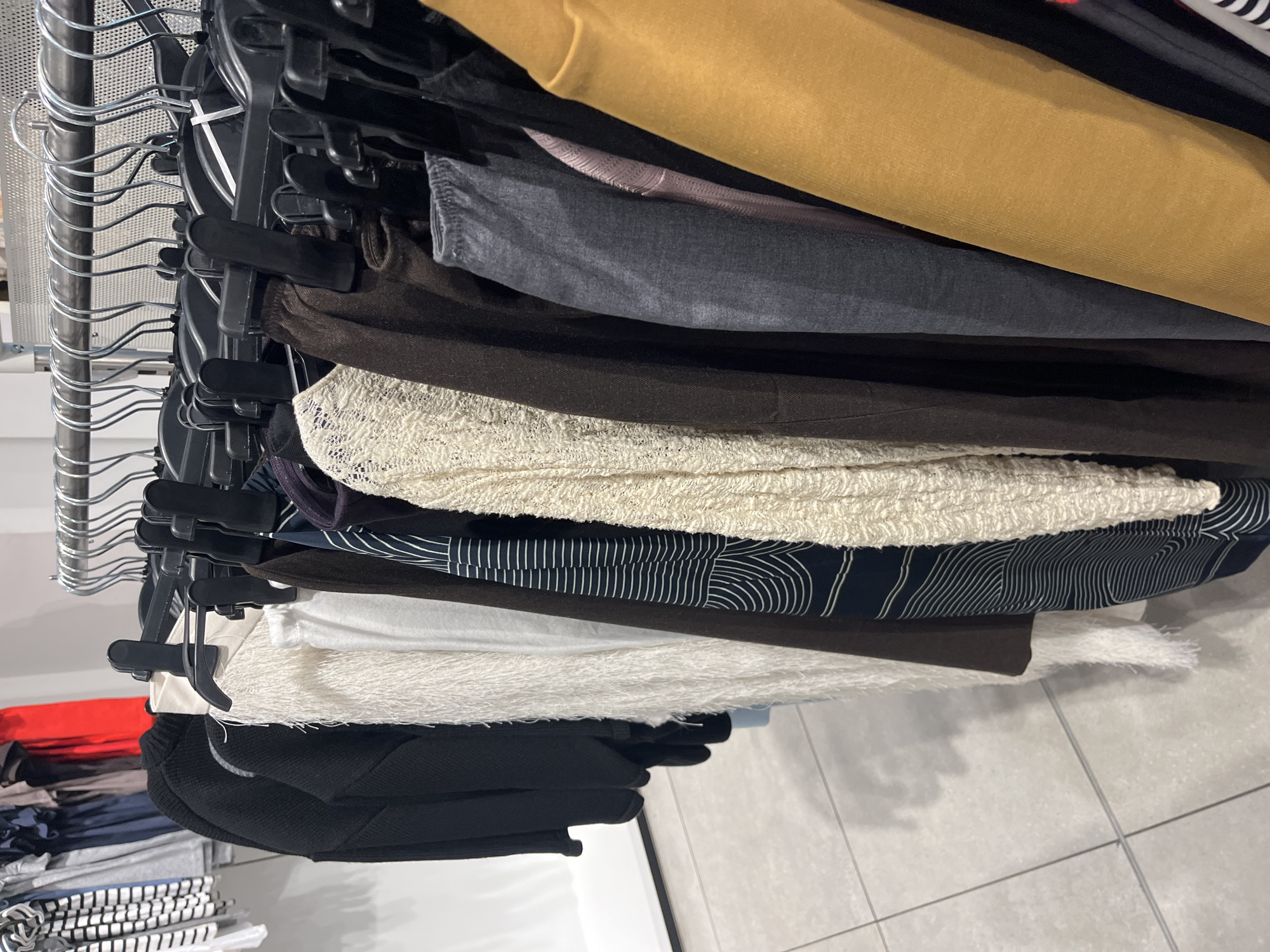 The image displays a close-up view of various clothing items neatly organized on a metal clothing rack, suggestive of a retail environment. A diverse selection of garments is visible, layered closely together. Dominant colors include a mustard yellow fabric at the top, followed by sections of dark grey, rich brown, and black materials. A cream or white textured fabric, possibly a knit or lace, is prominent among the layers. Below this, a dark blue garment with a distinctive white, wavy, abstract line pattern can be seen.
The clothes are held by black plastic hangers, which are suspended from a silver-colored metal rack with a series of curved supports, partially visible on the left side of the frame. The background features a light-colored tiled floor beneath the hanging garments and a plain light wall behind them. No people or visible text are present in the image. The scene conveys an organized display, likely within a clothing store located in Blagoevgrad, Bulgaria. The image is rotated 90 degrees clockwise from its natural orientation.