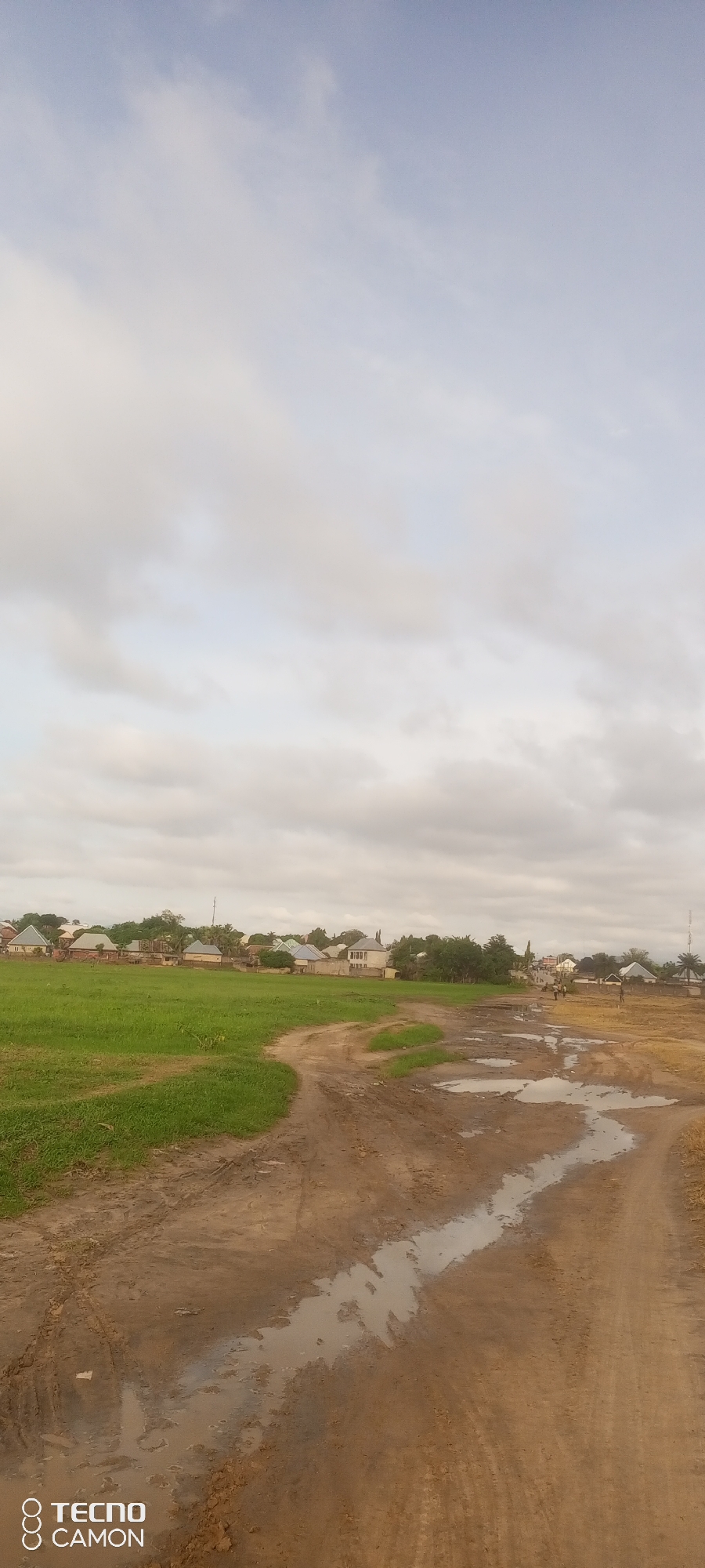The image depicts an overcast sky over a rural-urban fringe in Jalingo, Nigeria. In the foreground, a muddy, unpaved road with several puddles stretches towards a line of houses and trees. To the left of the road is a green, grassy field. The houses are a mix of styles, with some appearing to be single-story structures and others multi-story buildings. Several trees are interspersed among the houses. The overall impression is that of a developing area where a settlement is encroaching on agricultural or undeveloped land. The sky is a pale blue with scattered white clouds. A logo for "TECNO CAMON" is visible in the bottom left corner of the image.