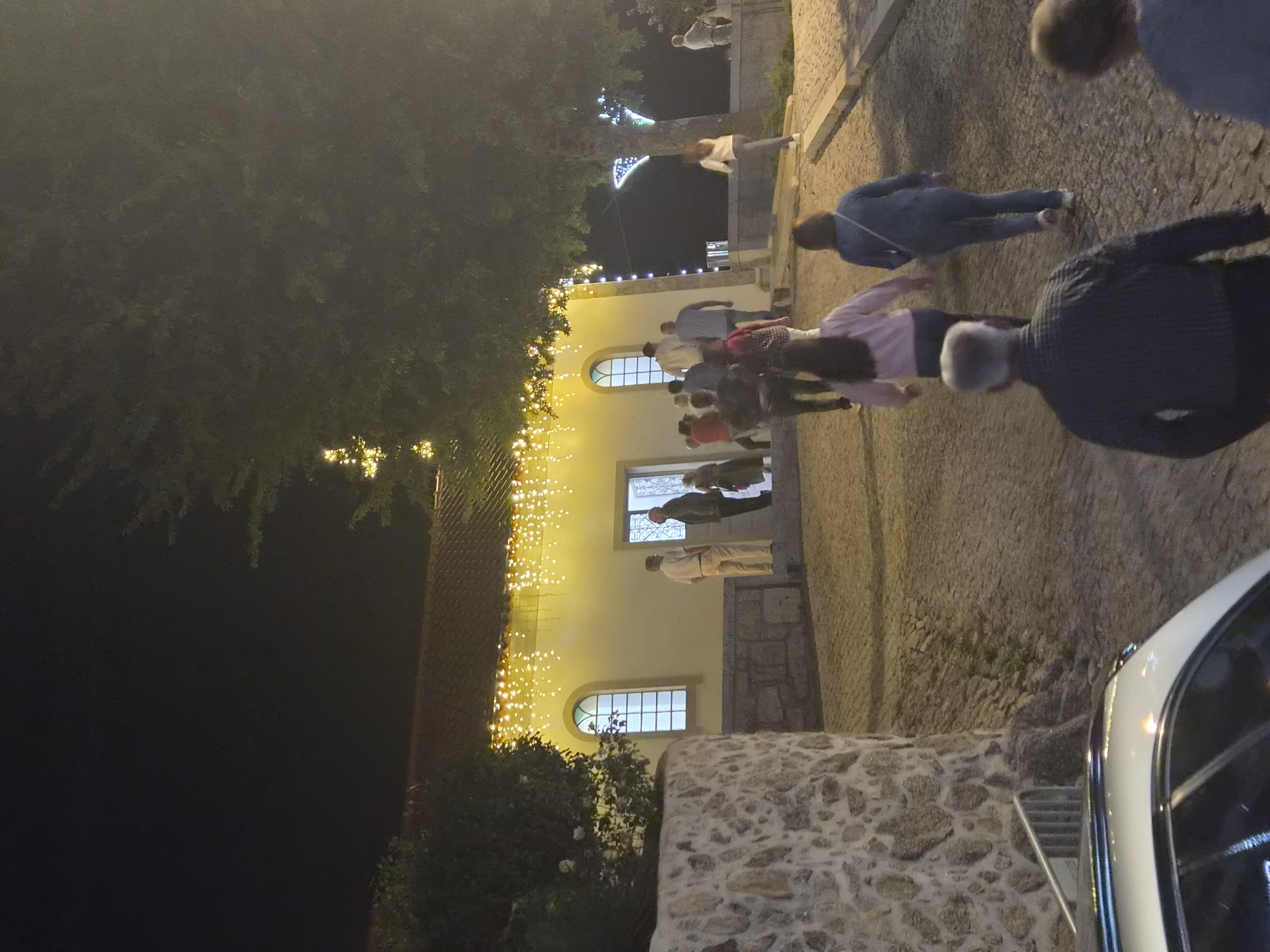 The image depicts a nighttime scene outside a building adorned with string lights. Several people are visible, some walking towards the entrance of the building, others standing near it. The building appears to be a church or a similar public structure, with arched windows and a pale yellow facade. The ground is paved with cobblestones. The overall atmosphere suggests a community gathering or event. The location is likely in Barcelos, Portugal, as indicated by the request.