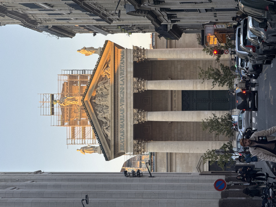 The image displays an urban street scene in Paris, France, centered on the Notre-Dame-de-Lorette church. The neoclassical church, situated at the end of the street, features a prominent portico with four large Corinthian columns, supporting a pediment adorned with bas-relief sculptures. Three statues are visible on the church's roofline, with the central and upper sections of the facade enveloped in extensive metal scaffolding, indicative of ongoing renovation. The inscription "BEATAE MARIAE VIRGINI LAVRETANAE" is visible above the columns, and "LIBERTE EGALITE FRATERNITE" is inscribed above the dark green main entrance doors.
The asphalt street in the foreground contains parked vehicles bumper-to-bumper along the right side. A black van is observed moving away from the viewer in the road, followed by a cyclist. In the immediate foreground, a man with a beard, wearing a light-colored jacket and a cap, walks towards the camera. Multiple pedestrians are distributed along both sidewalks, with a concentration visible on the left. To the left, a tall building with vertical fluting rises, while on the right, a multi-story Parisian-style building with balconies houses a ground-floor commercial establishment featuring a lit sign partially obscured but appearing to read "TABAC." A red traffic light is active on the right side of the street, and a circular red 'no parking/stopping' sign is mounted on a pole on the left. The upper portions of the church and its scaffolding are illuminated by warm, golden sunlight, indicating either early morning or late afternoon, while the street and lower structures remain largely in shadow.