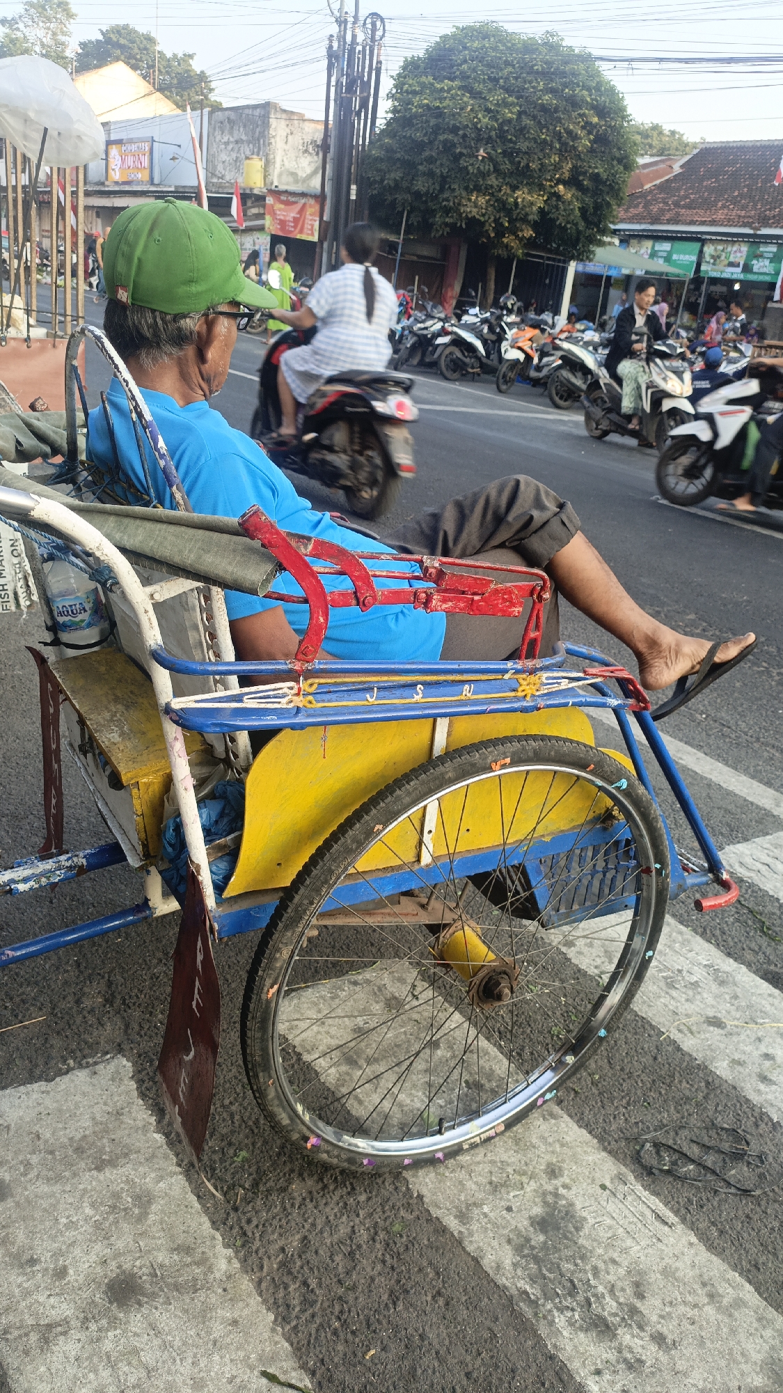 An elderly man wearing a green cap and a blue shirt is seated in a pedicab on a paved road in Srono, Indonesia. He is facing away from the camera. The pedicab has a yellow and blue frame with red accents. The man's legs are visible, and he is wearing dark pants and flip-flops. A bottle of Aqua water is attached to the side of the pedicab. In the background, there are several motorcycles parked and people walking. A large tree is visible on the right side of the image. The scene appears to be a street or market area in a small town.