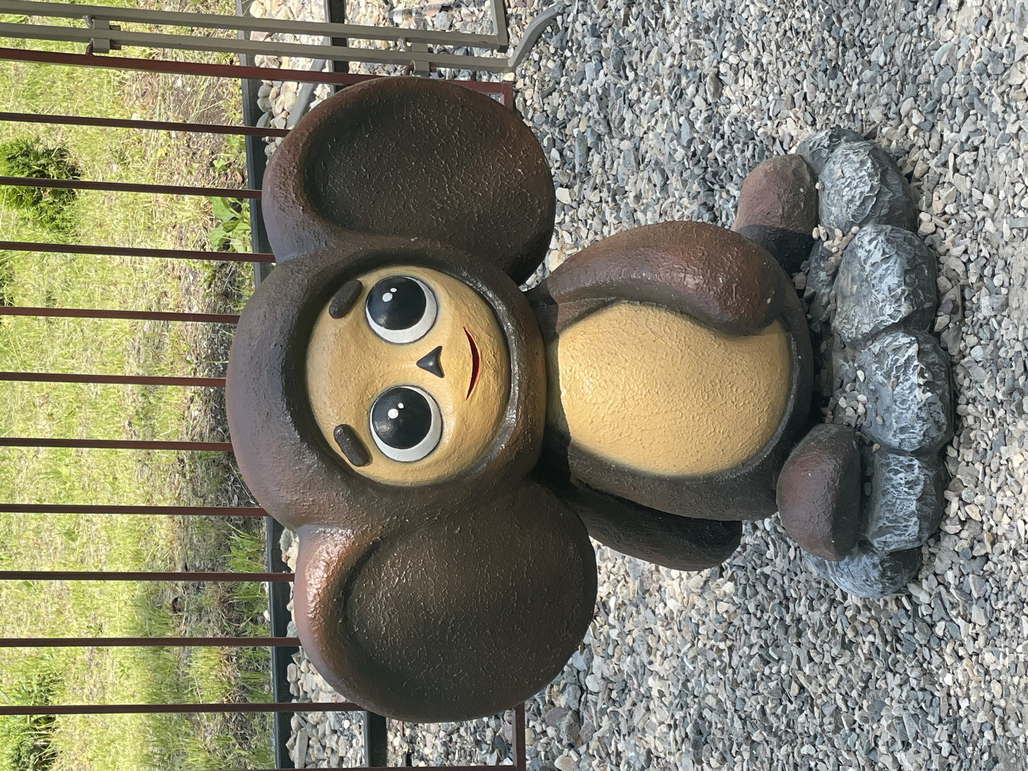 A statue of Cheburashka, a popular Russian cartoon character, sits on a bed of gravel. The statue is brown and tan, with large ears and big, round eyes. It is sitting on a base made of grey, rough-textured rocks, with small pebbles scattered around. Behind the statue is a metal fence with vertical bars, and beyond that, green grass and some patchy soil. The scene appears to be outdoors, possibly in a park or public area. The statue is centered in the frame, with the fence and grassy area forming the background.