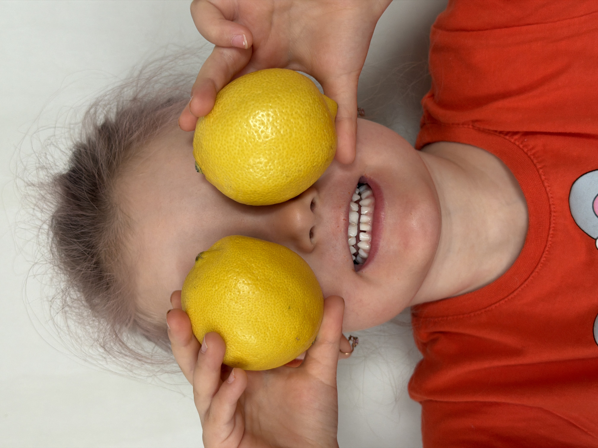 A child lies on their back, holding two lemons in front of their eyes. The child is fair-skinned, with blonde hair. They are wearing an orange shirt with a cartoon character on it. Their mouth is open, revealing their teeth. The background is white. The location is Chaikovsky, Russia.
