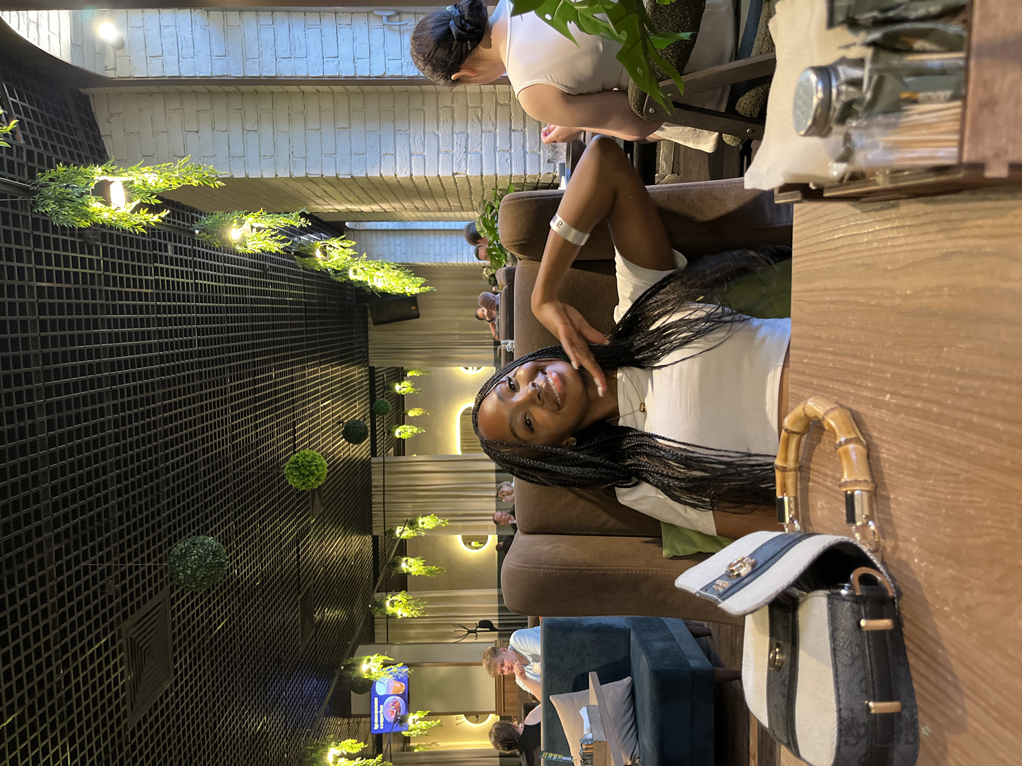 A young Black woman with long braids smiles at the camera, resting her chin on her hand. She is wearing a white t-shirt and sitting in a brown plush booth in a dimly lit restaurant with hanging plants and a black, grid-like ceiling. To her left, a stylish handbag with a bamboo handle rests on a wooden table. In the background, other patrons are visible, some dining, some conversing. The restaurant appears to be modern and decorated with greenery. The scene suggests a relaxed dining experience.