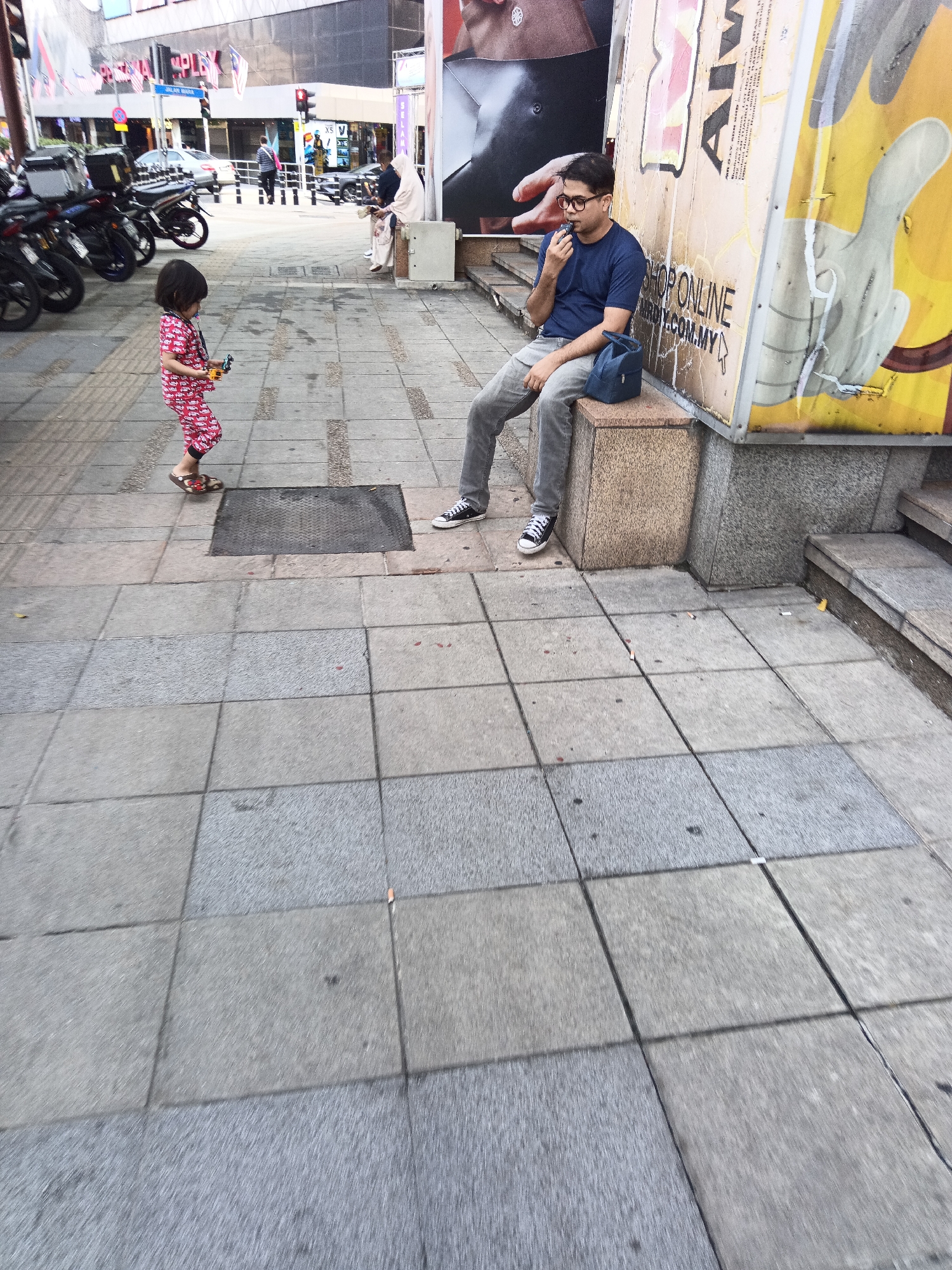 A young child in red patterned pajamas and sandals stands on a tiled sidewalk, holding a small toy. Nearby, a man wearing a blue t-shirt, grey pants, and glasses sits on a stone bench, appearing to eat or vape. He has a blue bag at his side. In the background, motorcycles are parked along the street, and a building with signage is visible. The scene takes place on a street in Kuala Lumpur, Malaysia, indicated by the "SHOP ONLINE" billboard in the background, a common marketing method in the city. The overall environment is urban, with pedestrians and commercial establishments.