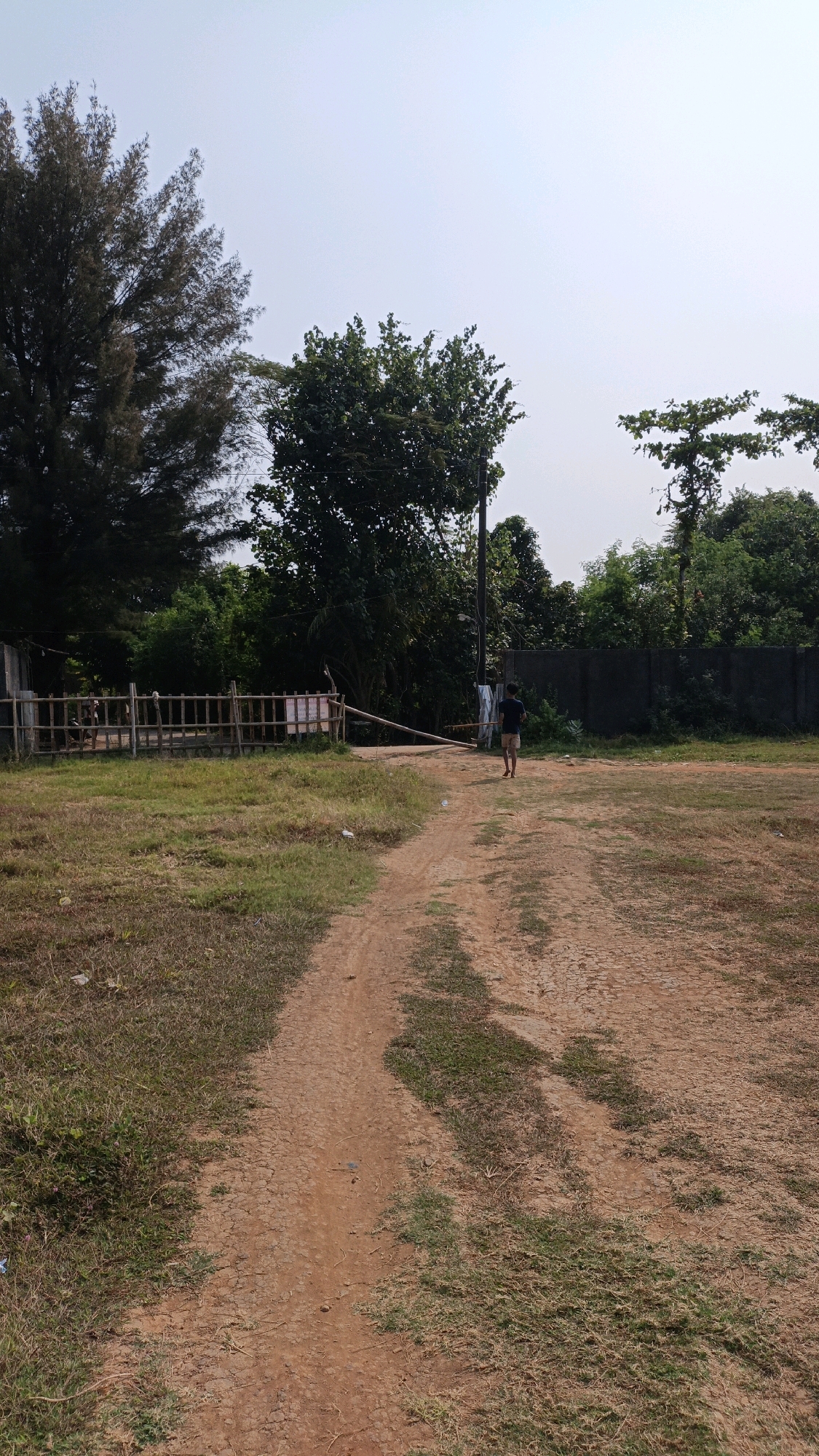 A solitary person walks away from the camera on a dirt path in an open, grassy area. The path is reddish-brown and appears to be a track made by vehicles. To the left of the path, there is a bamboo fence surrounding an area with some scattered debris and a building. Beyond the fence, tall trees with dark foliage are visible. To the right of the path, the ground is mostly dry grass and dirt. In the background, more trees and a concrete wall can be seen, with a utility pole with a light fixture standing near the middle of the frame. The sky is a pale, hazy blue. The image appears to be taken in Cikal, Indonesia, likely in an undeveloped or recreational area, possibly a park or a field on the outskirts of the city.
