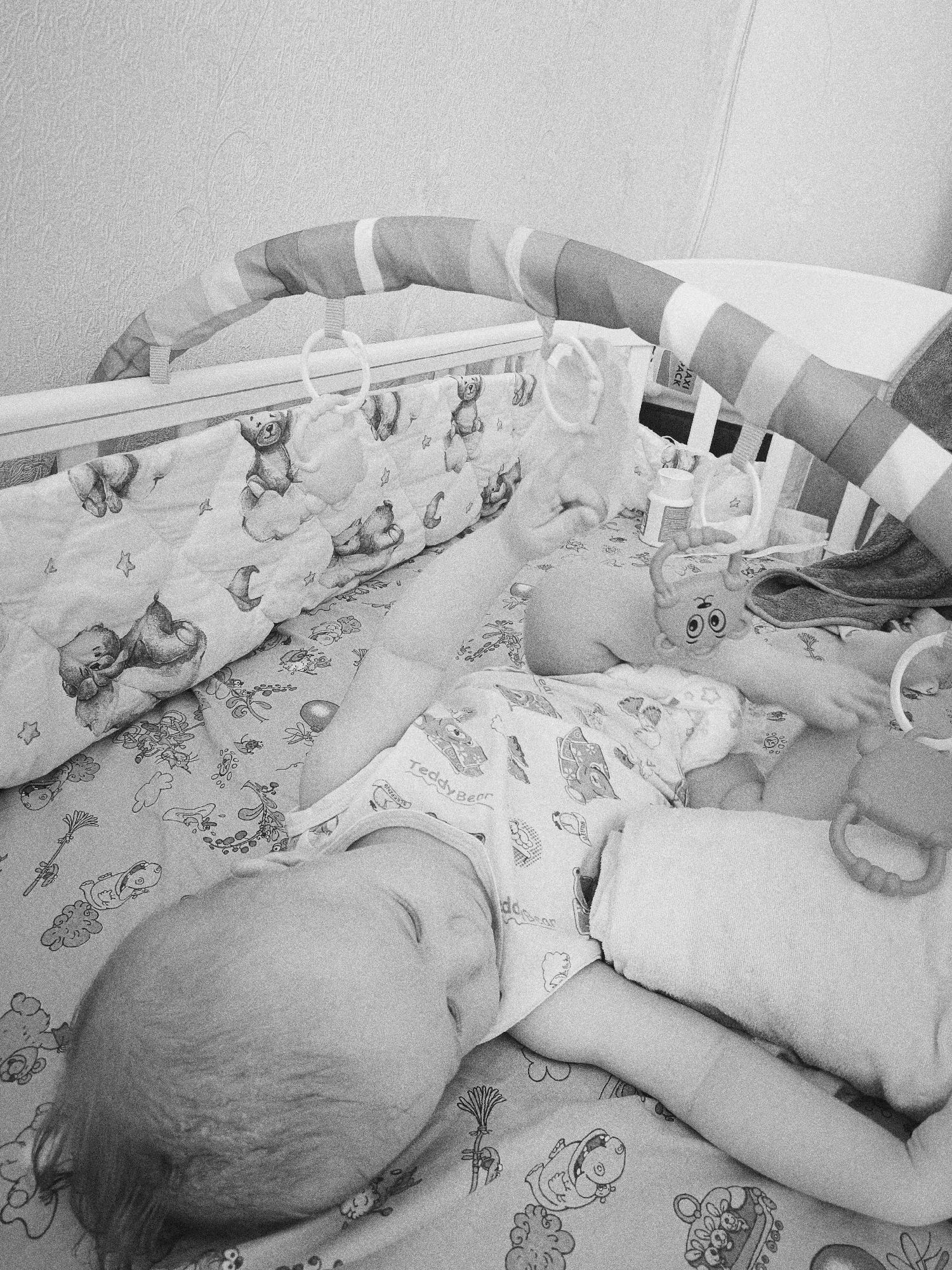 A black and white photograph depicts a baby sleeping in a crib. The baby is lying on a patterned sheet with its head turned to the left. The baby's eyes are closed. The baby is wearing a patterned onesie. A crib mobile hangs above the baby. The crib has padded sides. The location is unknown.